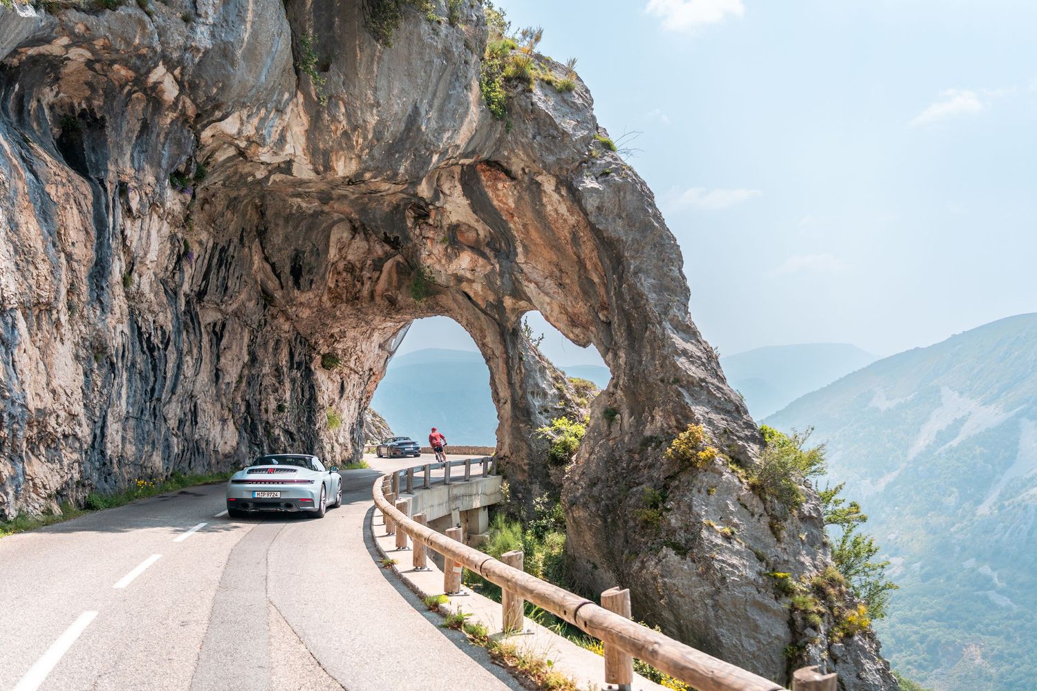 A silver sports car drives through a scenic mountain tunnel carved into the rock along a winding coastal road.
