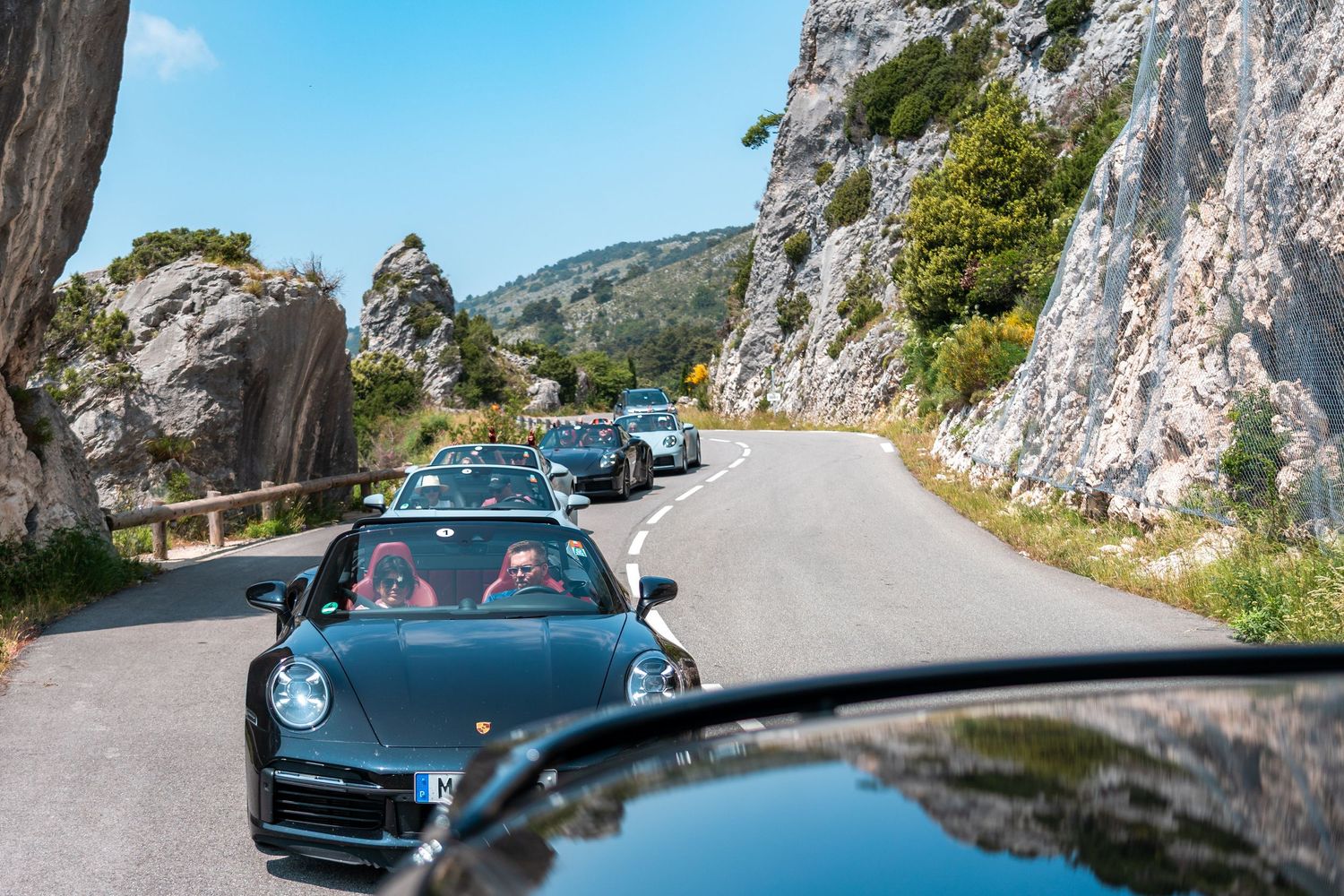 A dark blue vintage Porsche sports car drives along a scenic mountain road with rocky cliffs and water views.
