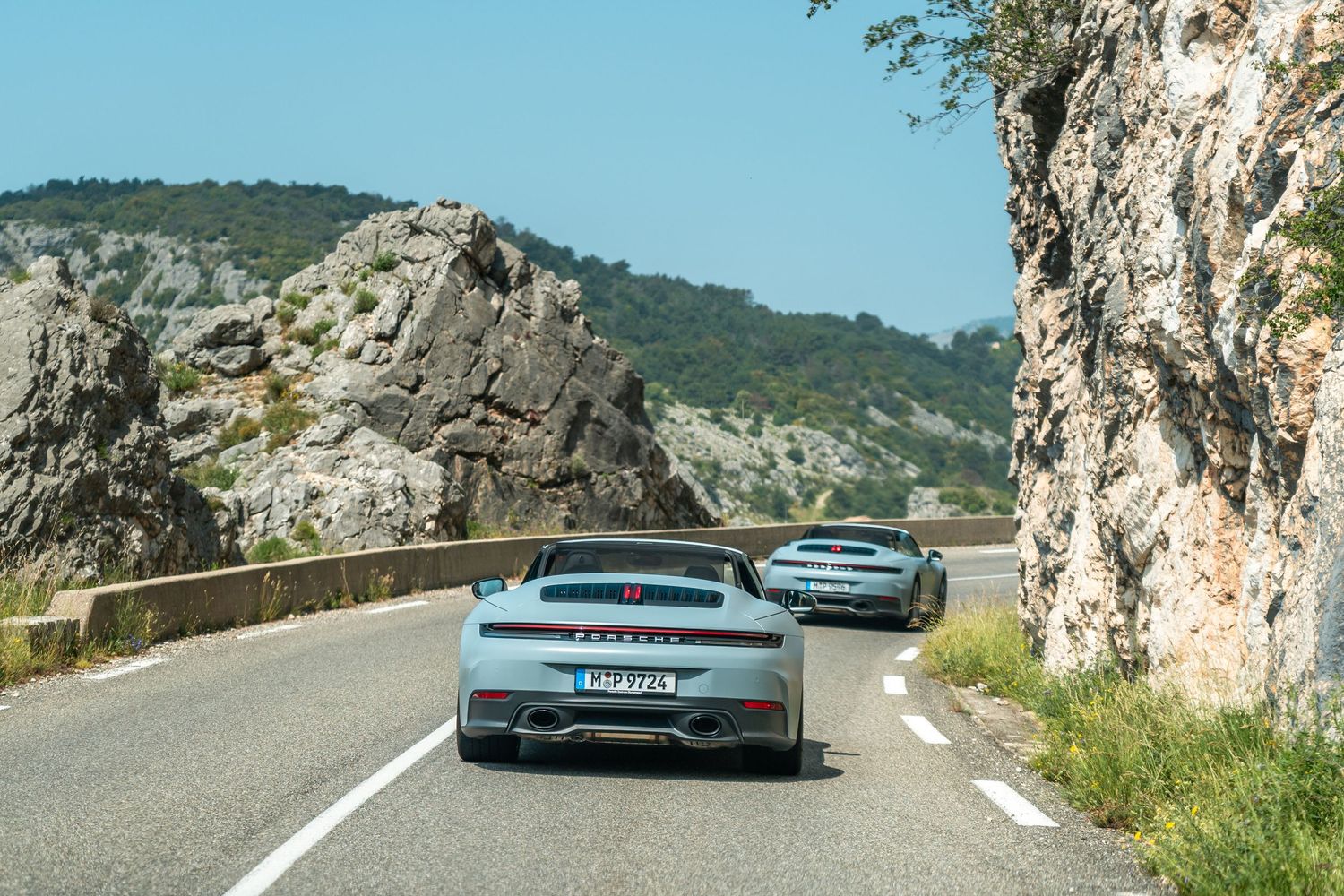 Two Porsche sports cars driving on a winding mountain road with rocky cliffs and scenic landscape views.
