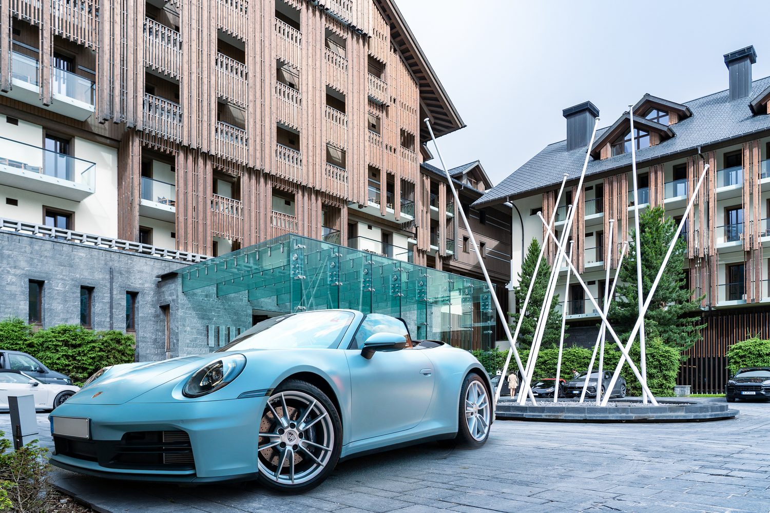 A light blue Porsche 911 parked in front of modern architectural buildings with sculptural elements.