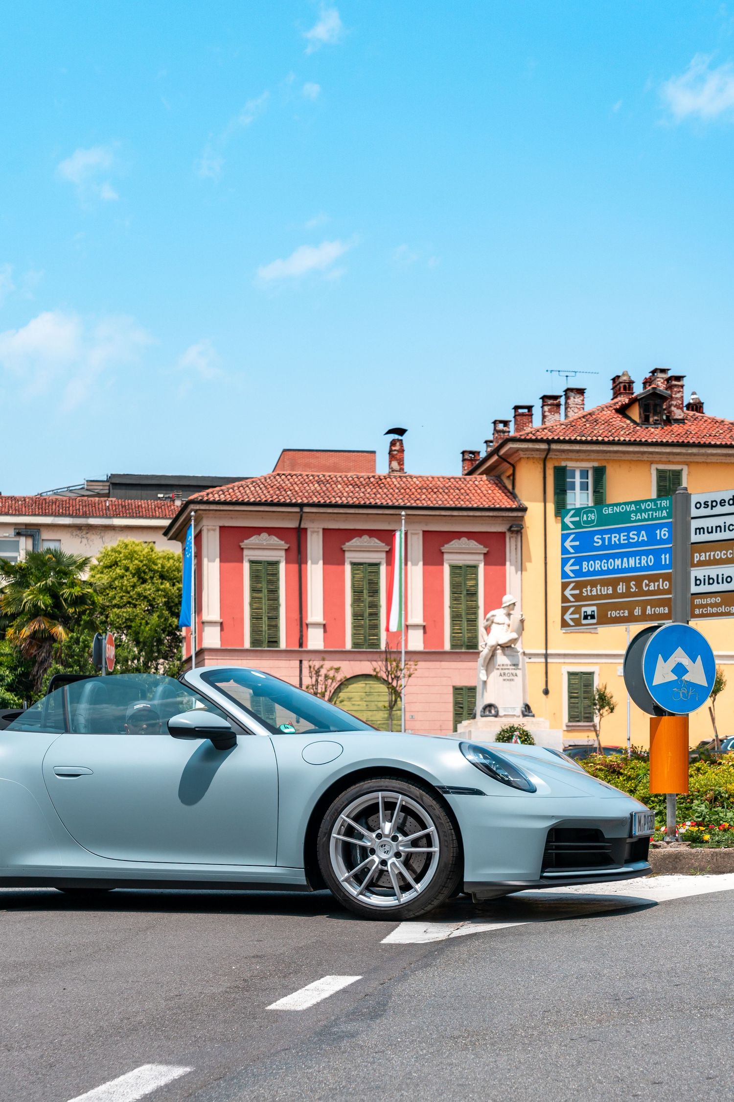A sleek silver Porsche 911 parked on a street with colorful Italian buildings and road signs in the background.