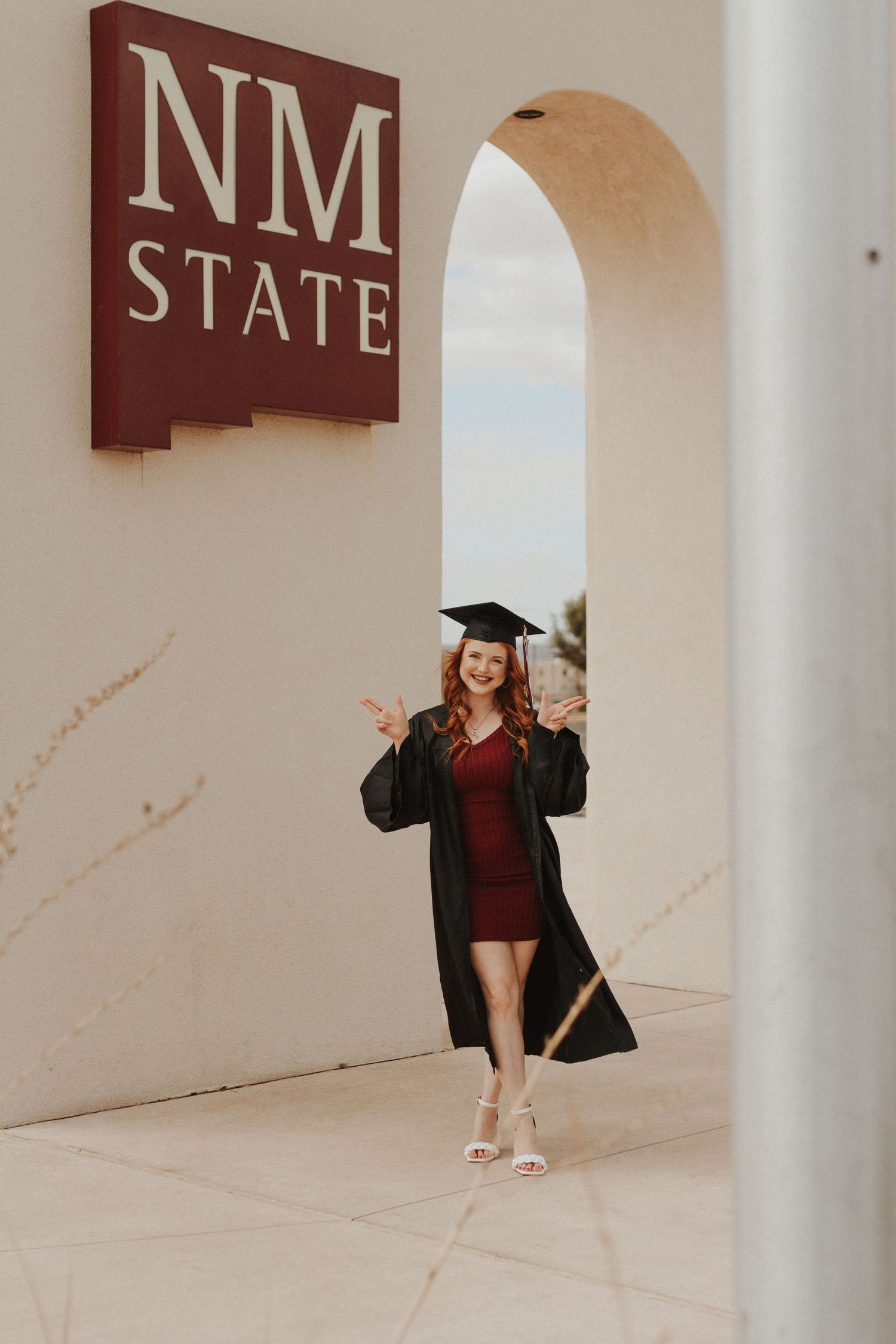 Graduate poses under NM State sign arch in traditional cap and gown.