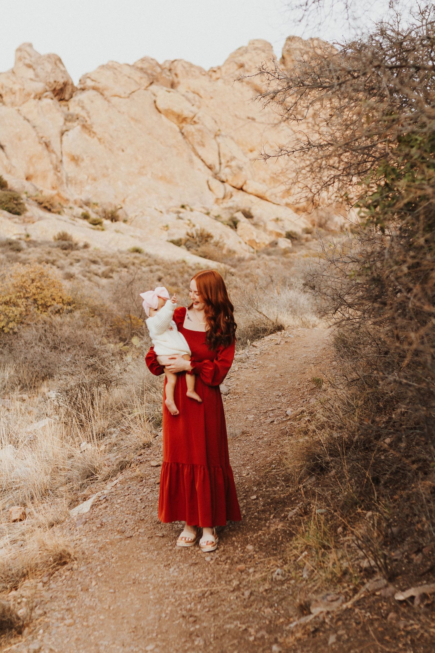 A mother in a flowing red dress holds her child on a desert hiking trail.
