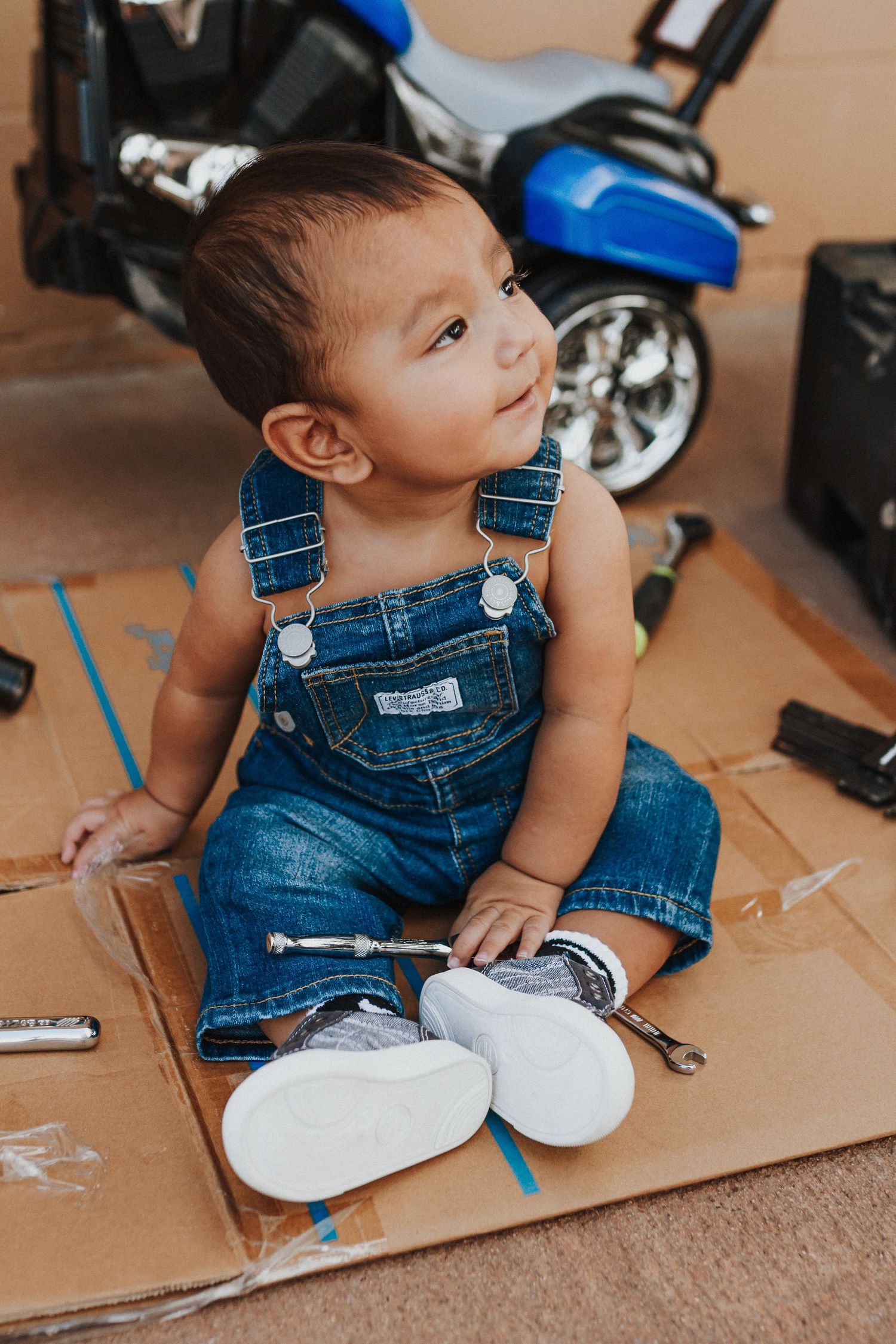 Baby in denim overalls sitting on cardboard playing with white shoes.