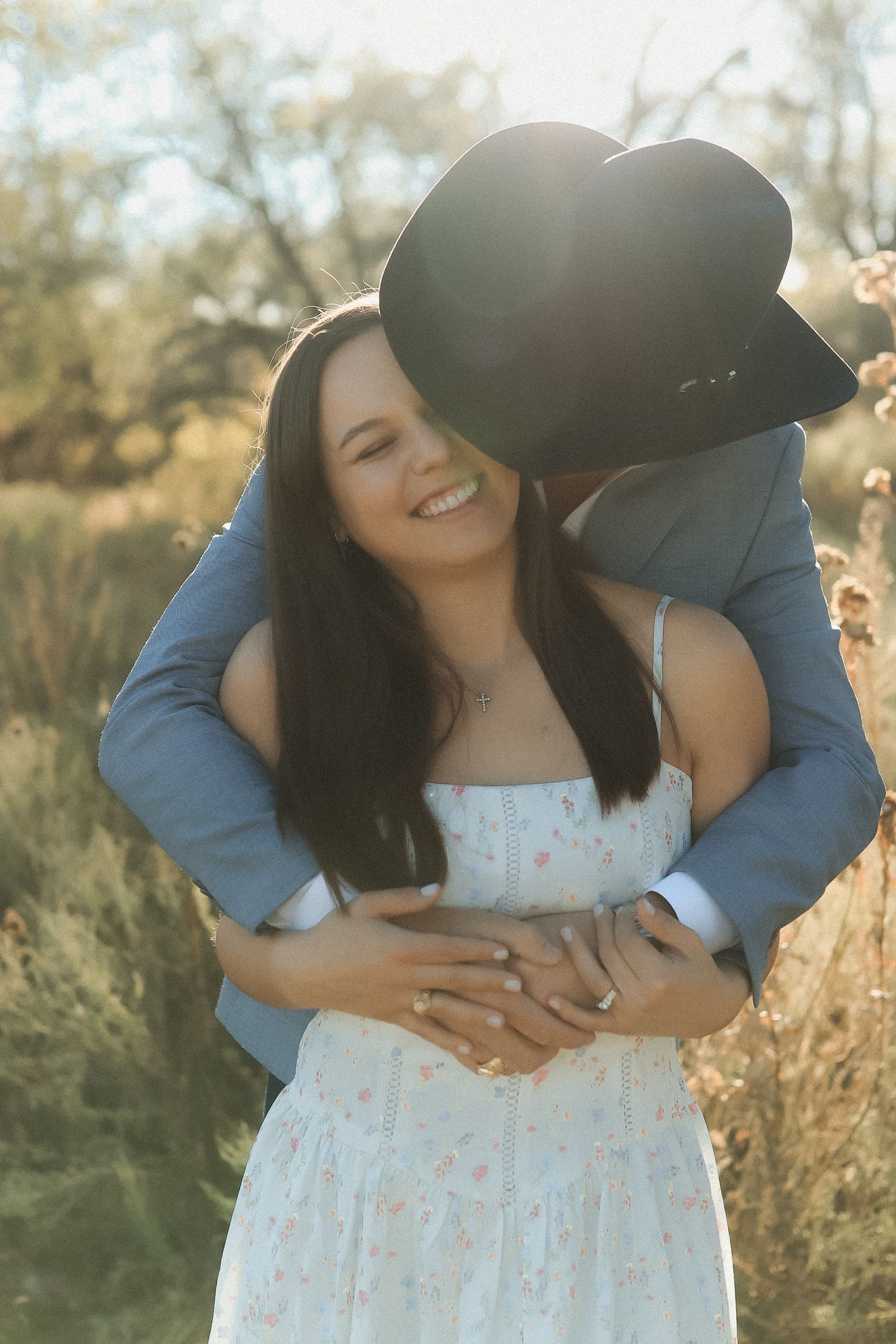 A smiling woman in a floral dress is embraced from behind by a man in a cowboy hat outdoors in golden light.