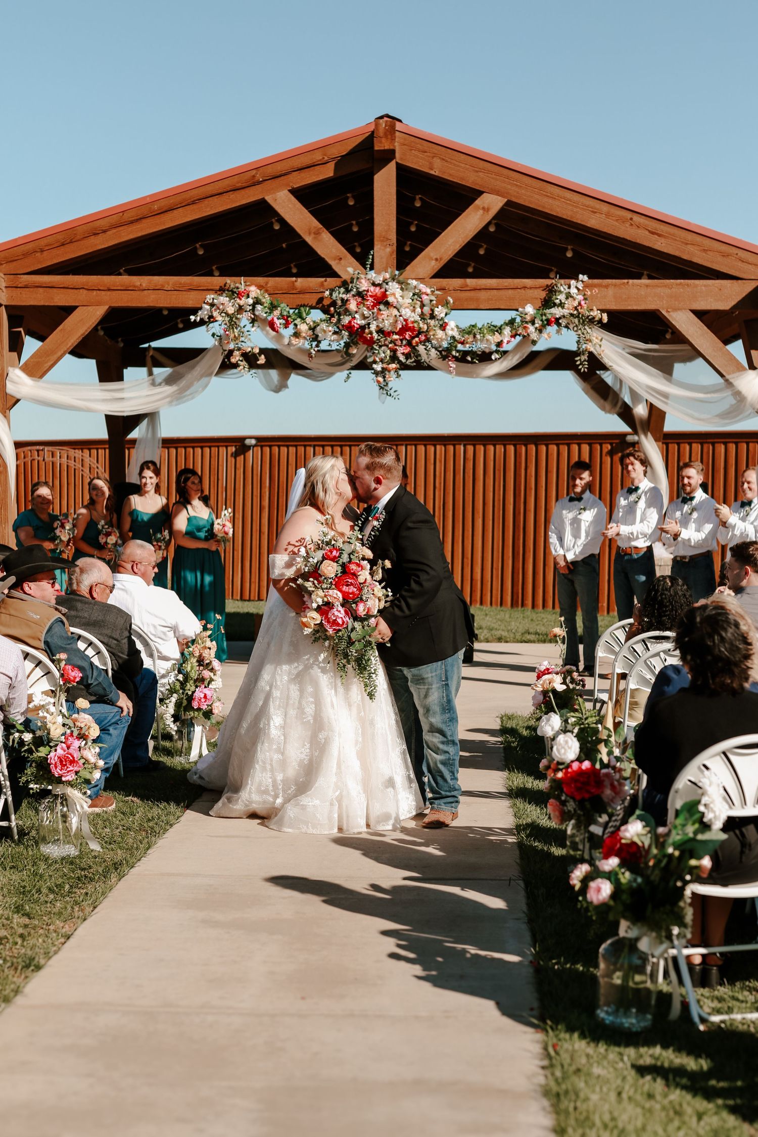 Bride and groom share first kiss at outdoor rustic wedding ceremony under wooden pavilion adorned with floral garlands.