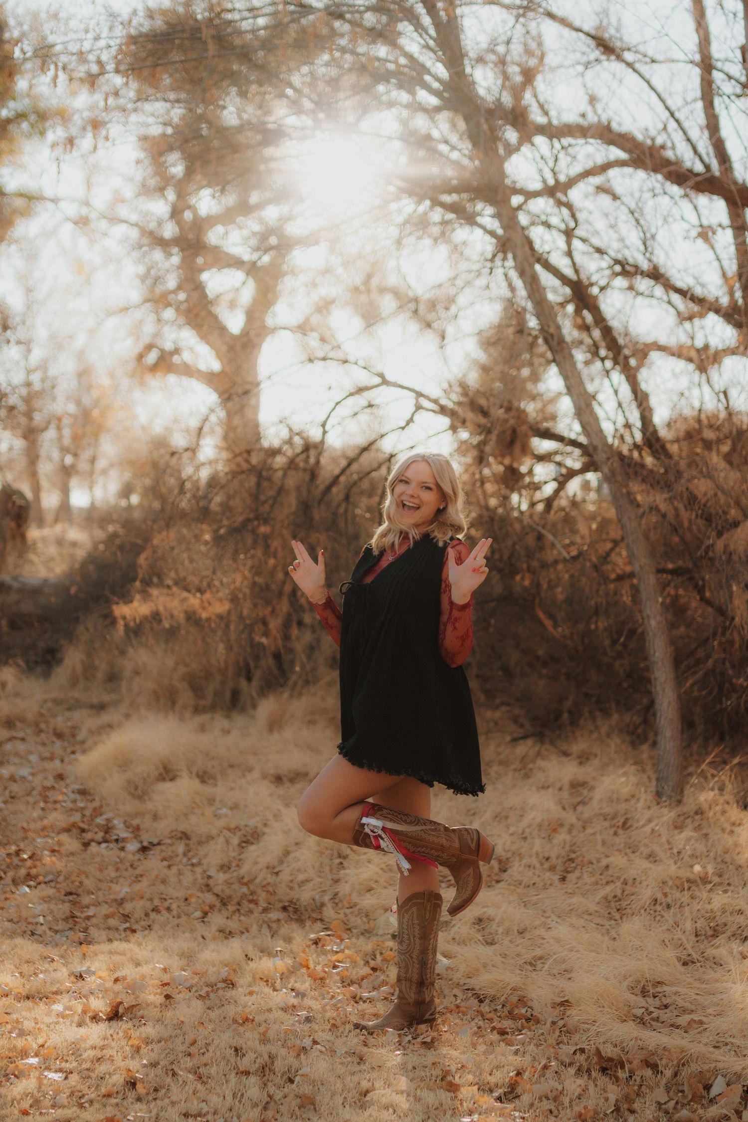 Playful graduate in cowboy boots poses against autumn trees in golden light.