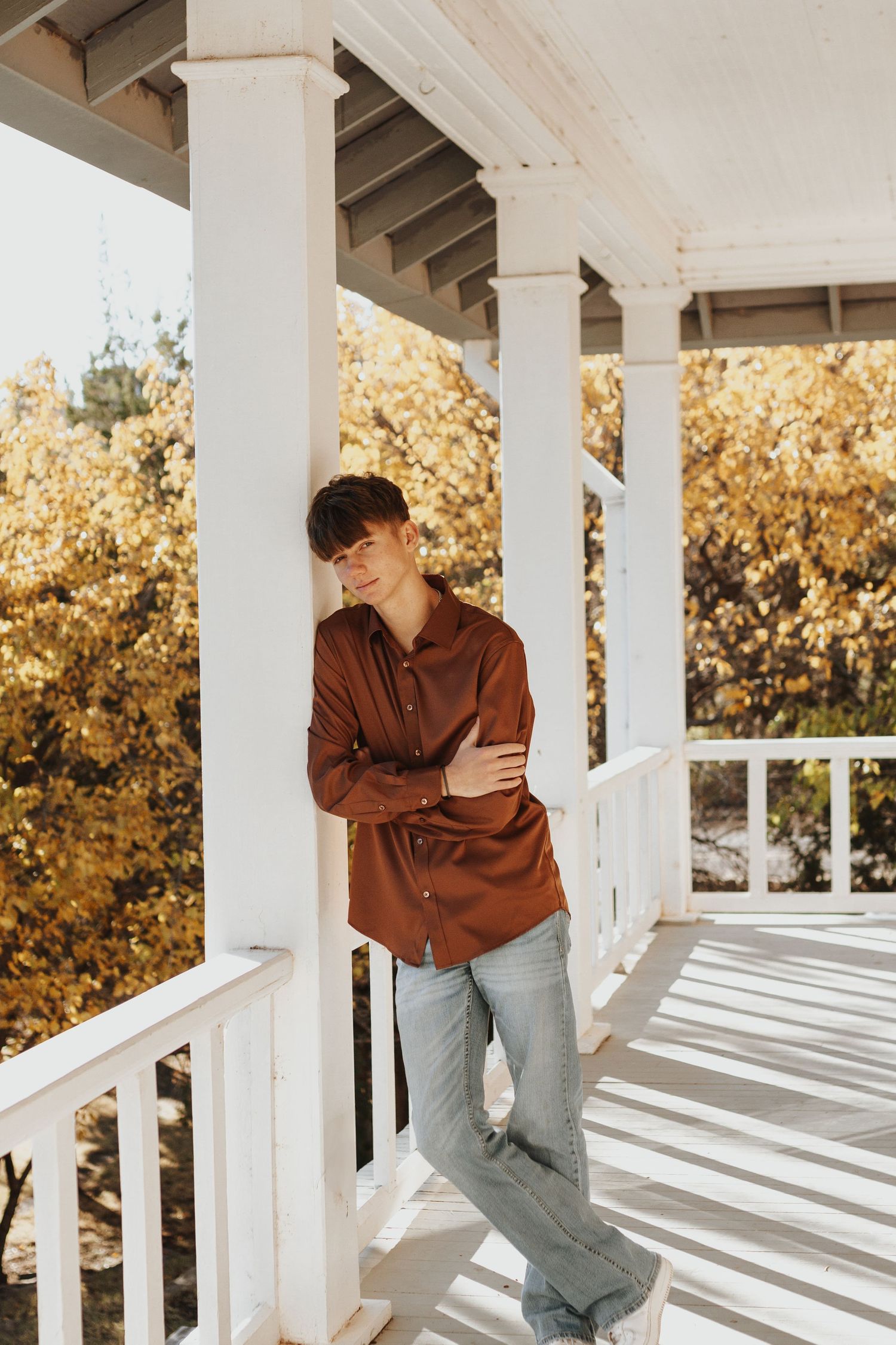 Person in brown shirt leaning against white porch pillars in autumn setting.