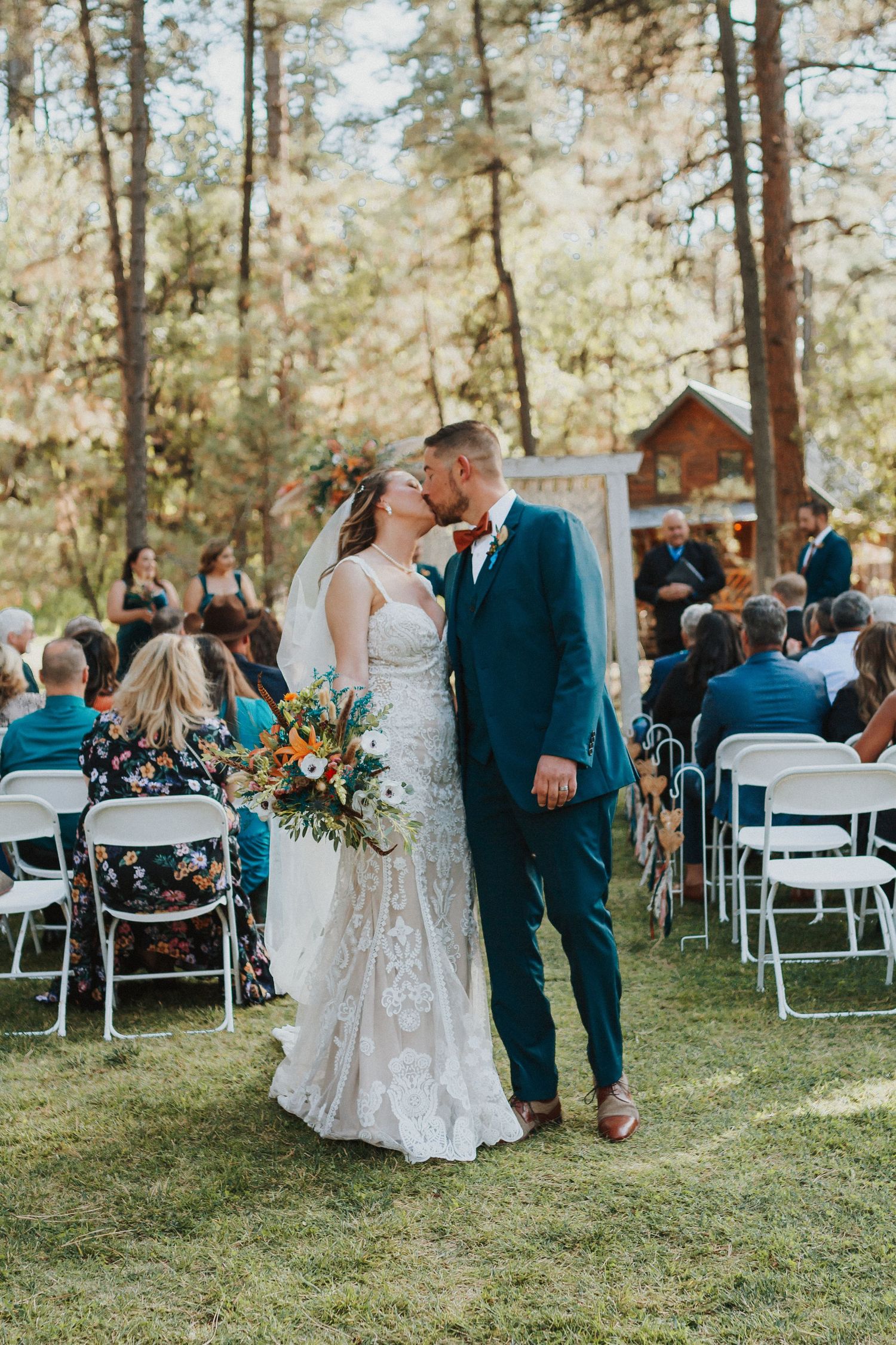 Outdoor forest wedding ceremony with white chairs and navy-suited guests.