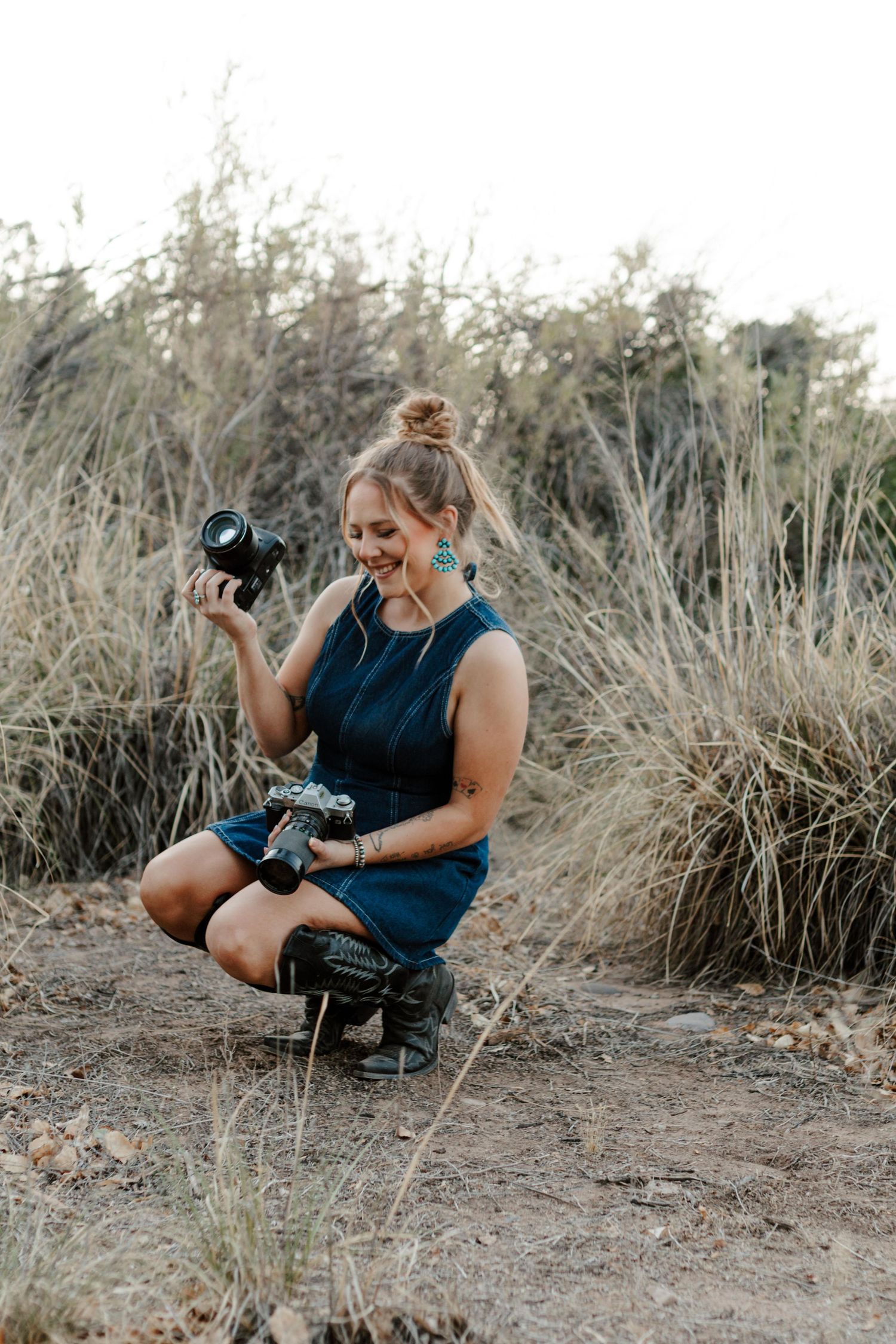 Woman photographer crouching outdoors in teal dress, smiling while holding DSLR camera among dry grasses.