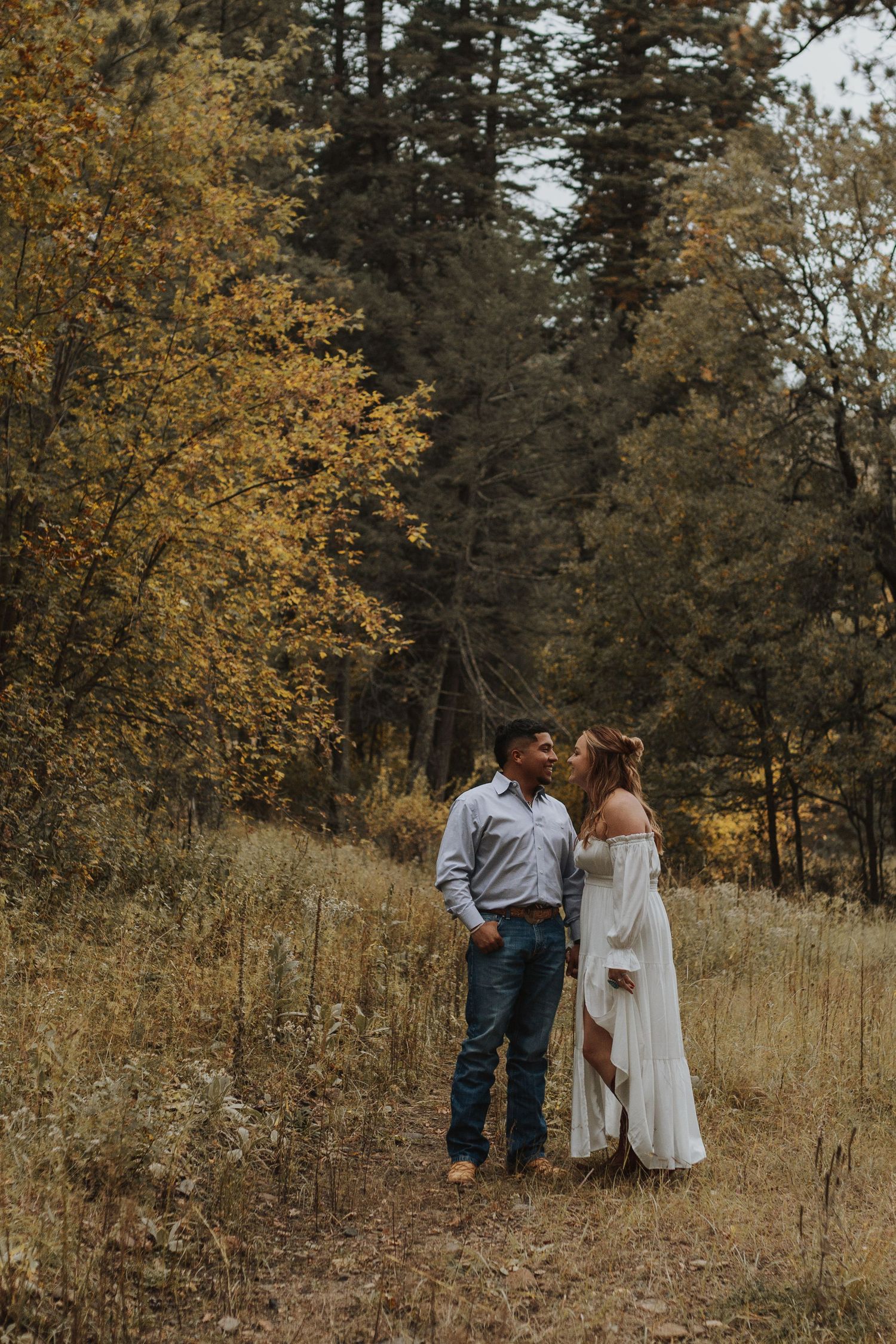 Couple sharing a kiss in an autumn forest clearing, woman in white dress and man in jeans surrounded by golden fall foliage.