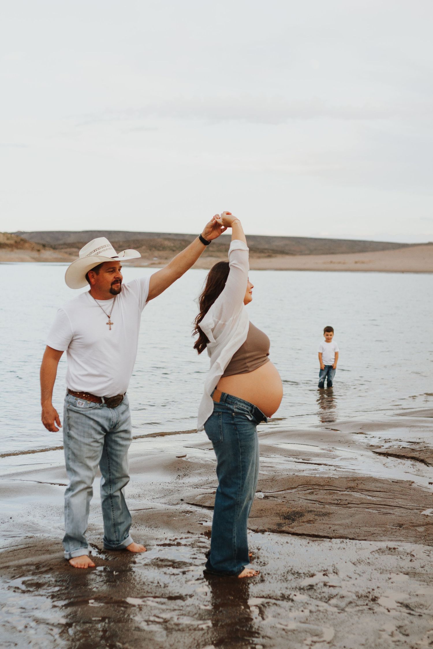 Family playing and dancing in shallow water at beach during overcast day.