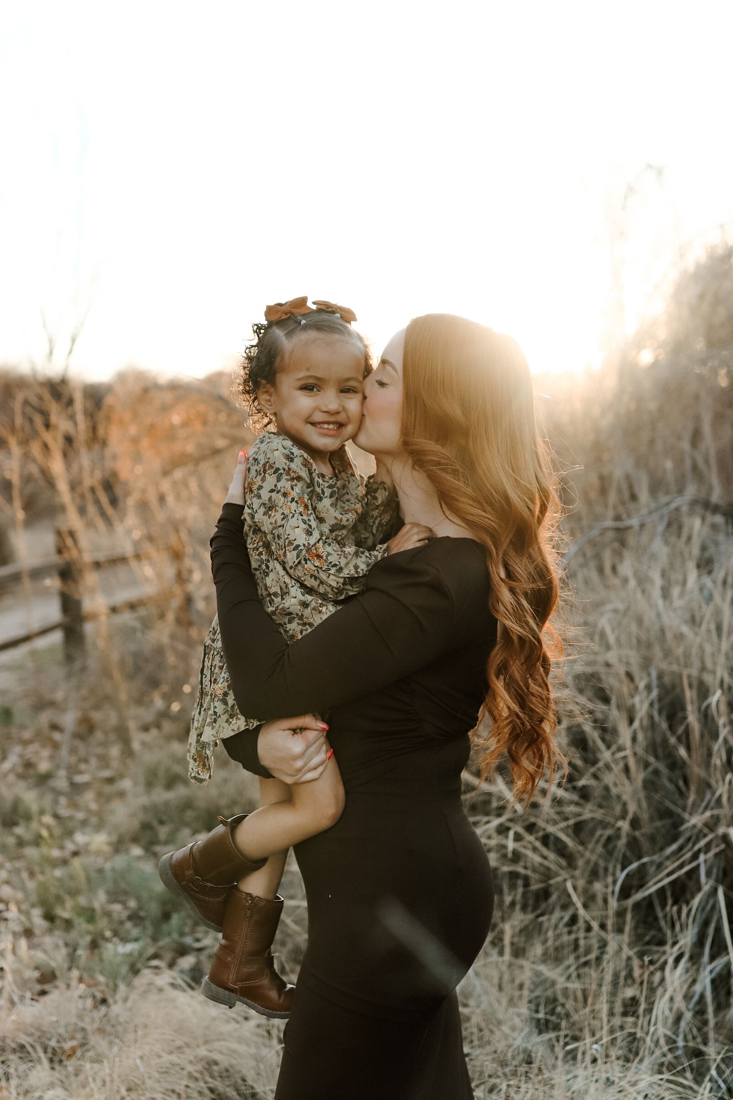 A mother with long wavy hair kisses her smiling toddler girl outdoors at golden hour sunset.
