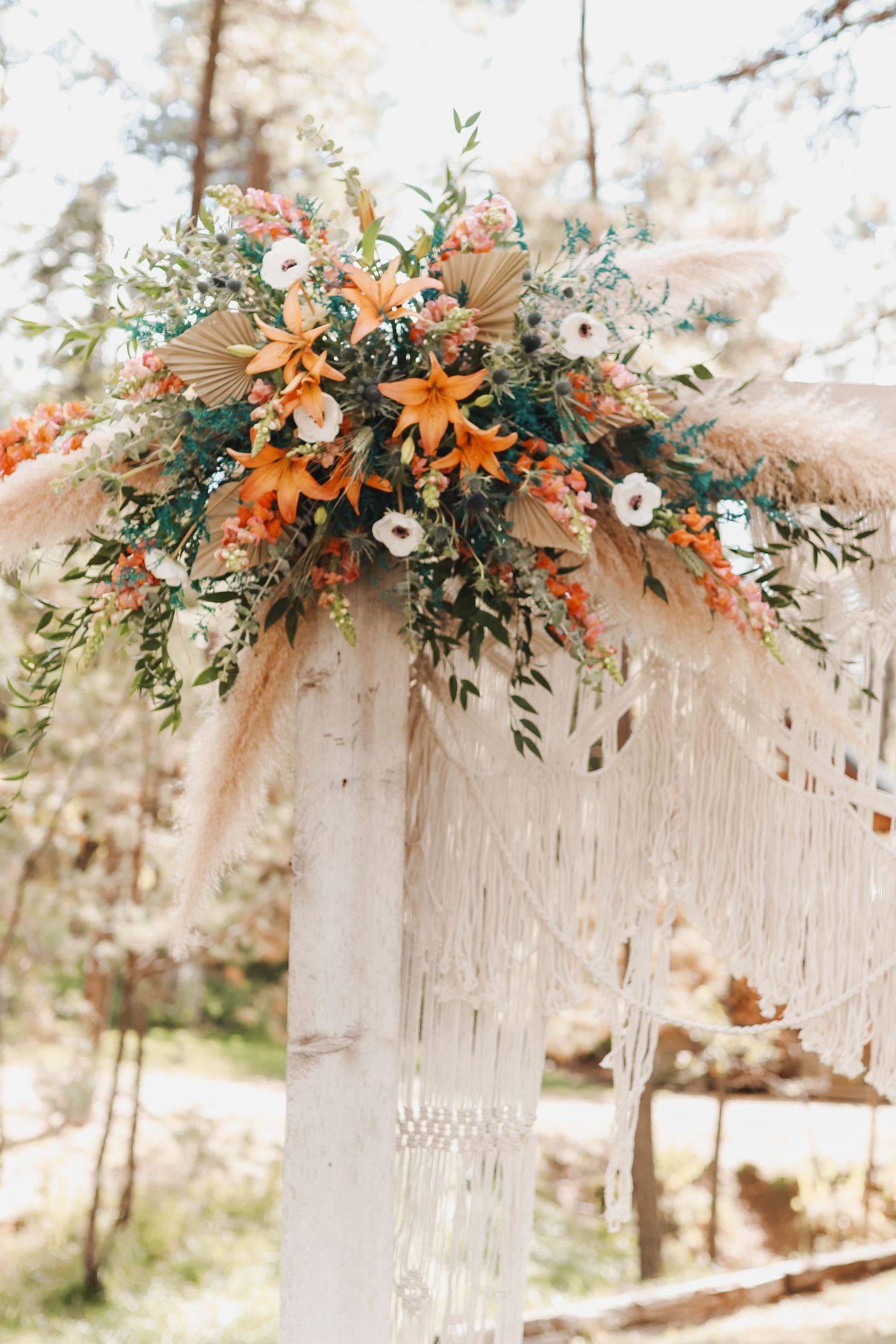 A rustic floral arrangement with orange lilies, pampas grass, and eucalyptus decorates a white wooden arch outdoors.