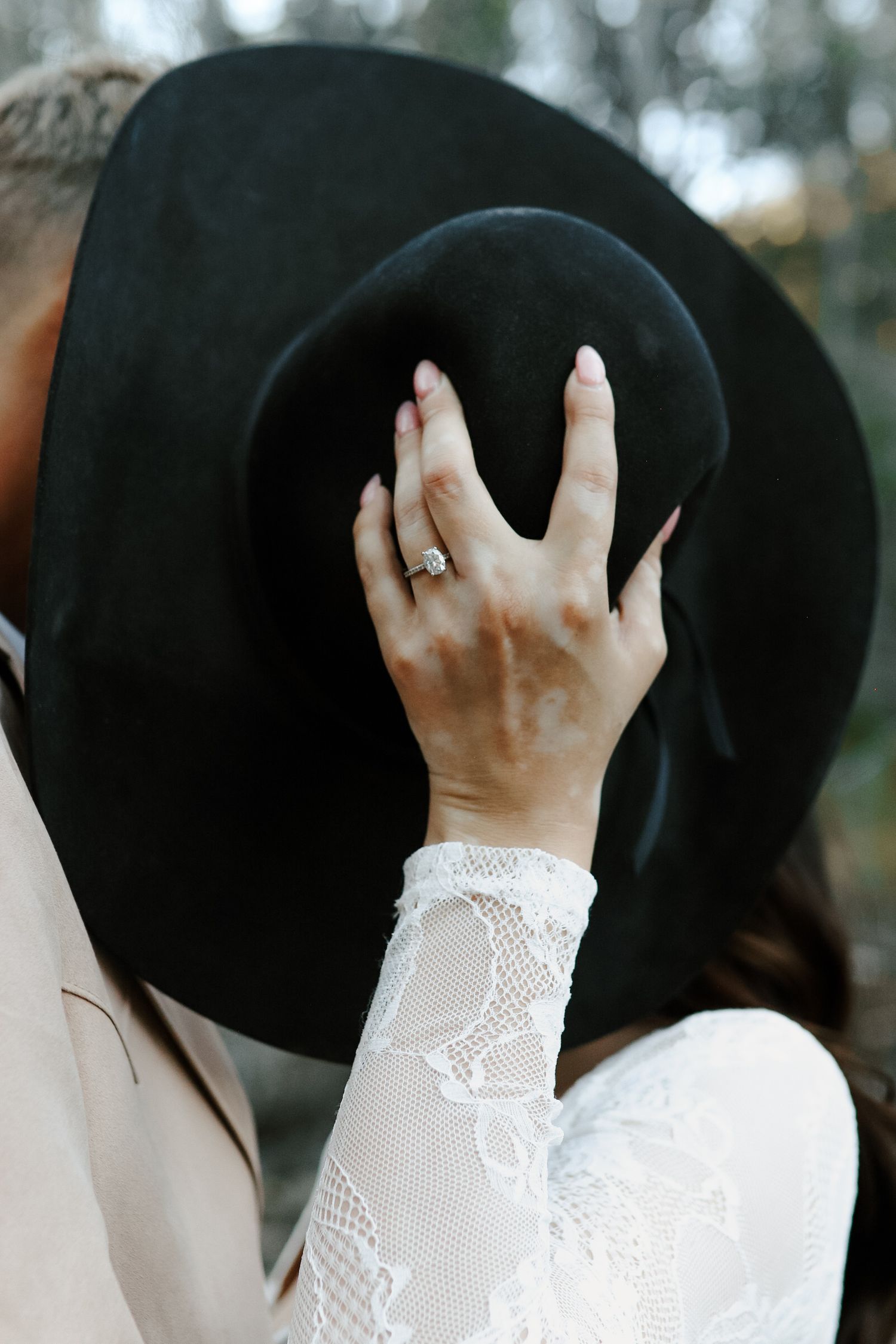 Close-up of a bride's hand holding a black wide-brim hat, showcasing a diamond engagement ring and lace sleeve.