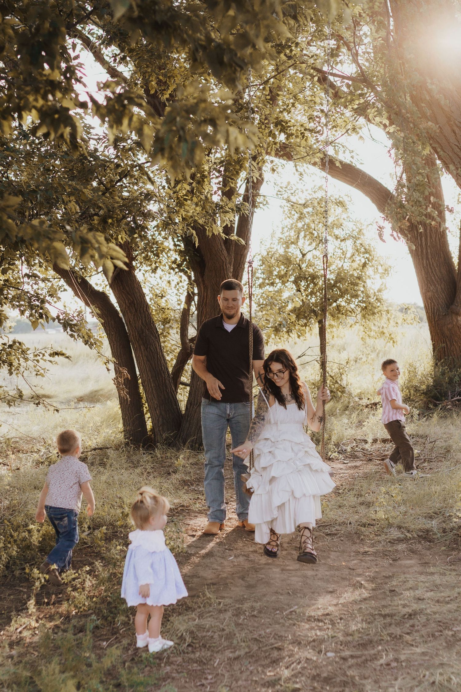 Family enjoying sunny outdoor time near large trees, mother in white dress on swing, father and children playing in nature.
