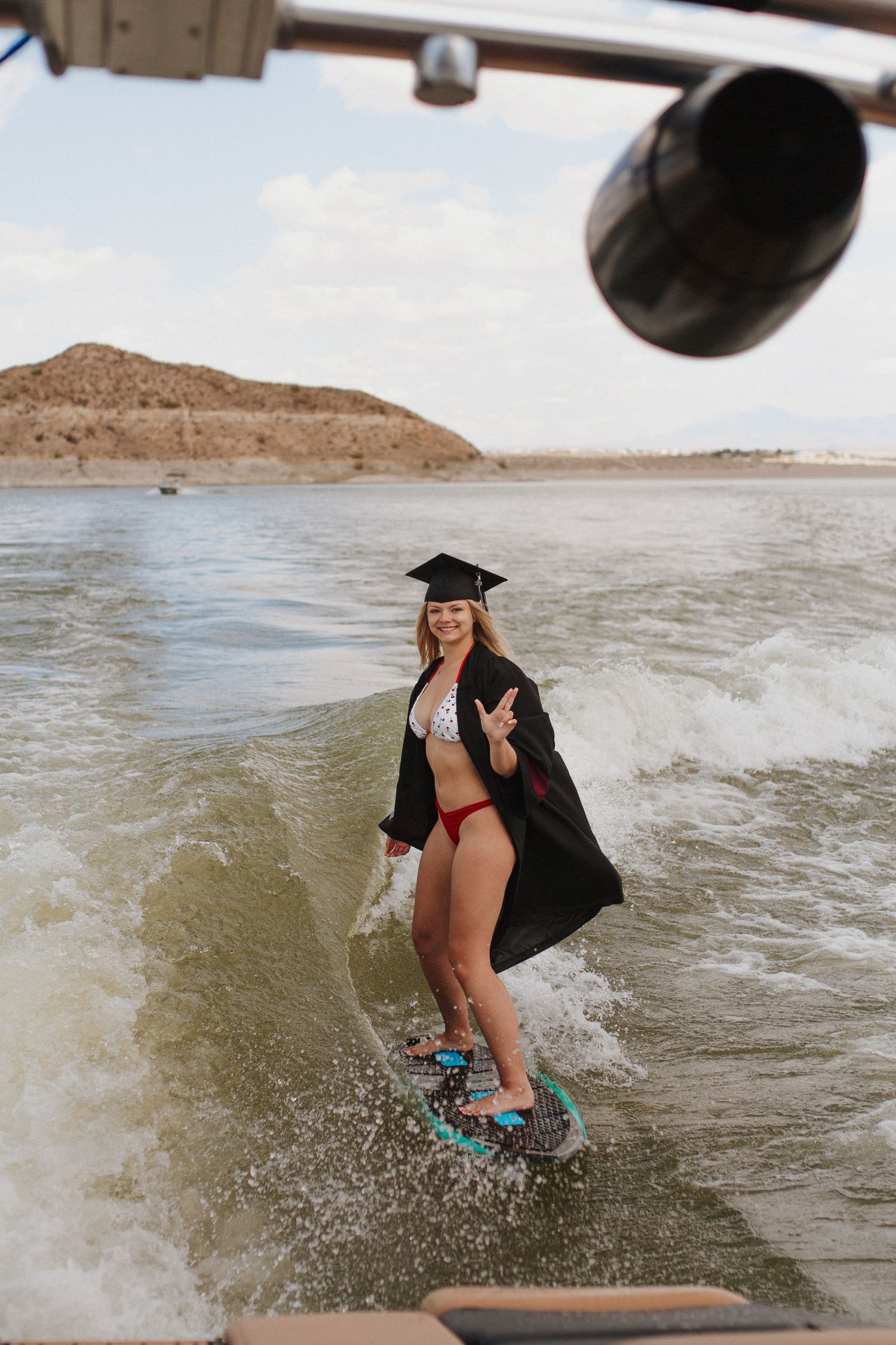 Graduate wakeboarding in cap and gown celebrates on lake during sunset photo session.