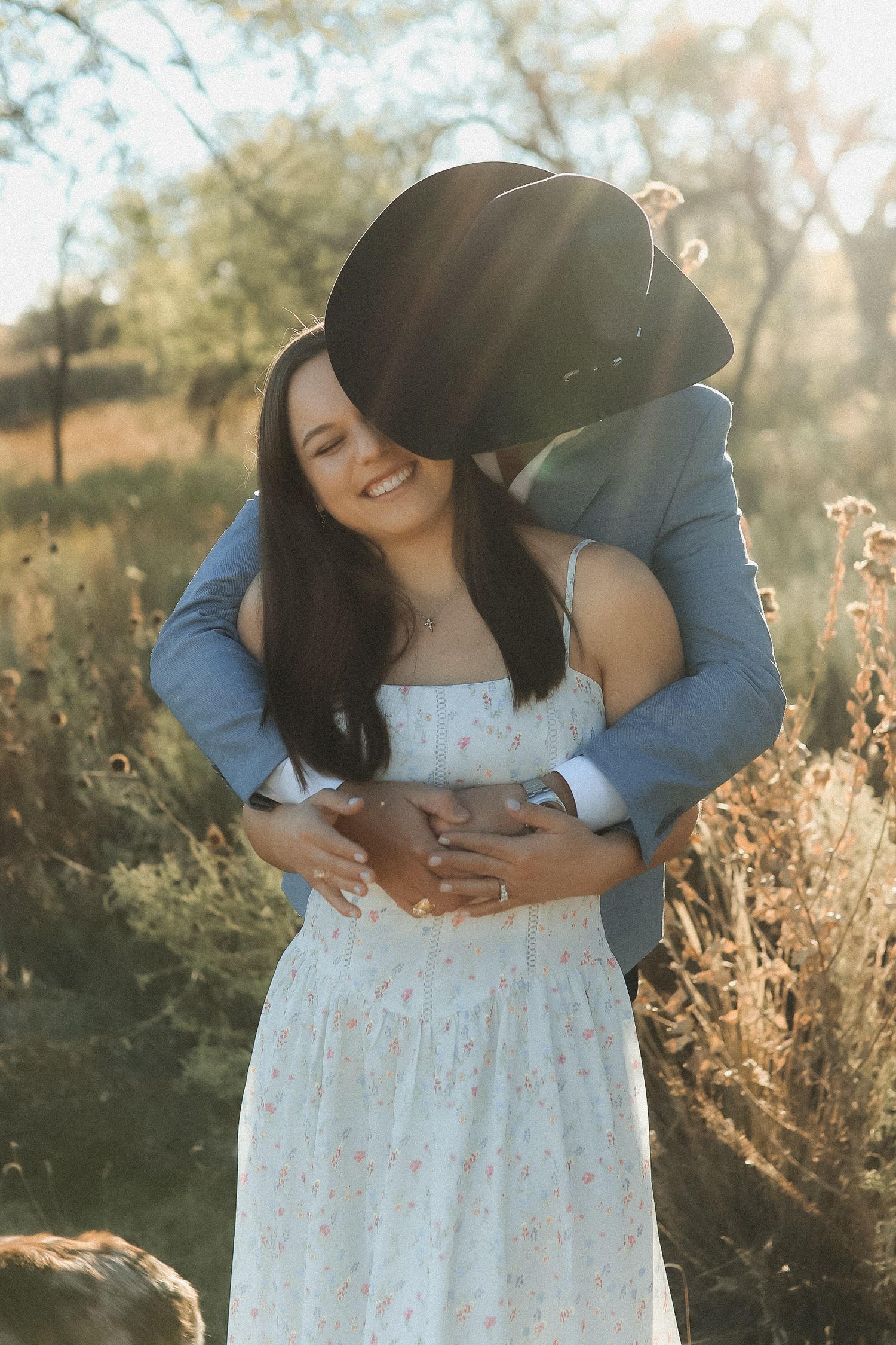 Couple embracing in golden sunlight, woman in floral dress smiling as man in blue suit and cowboy hat hugs her in a field.