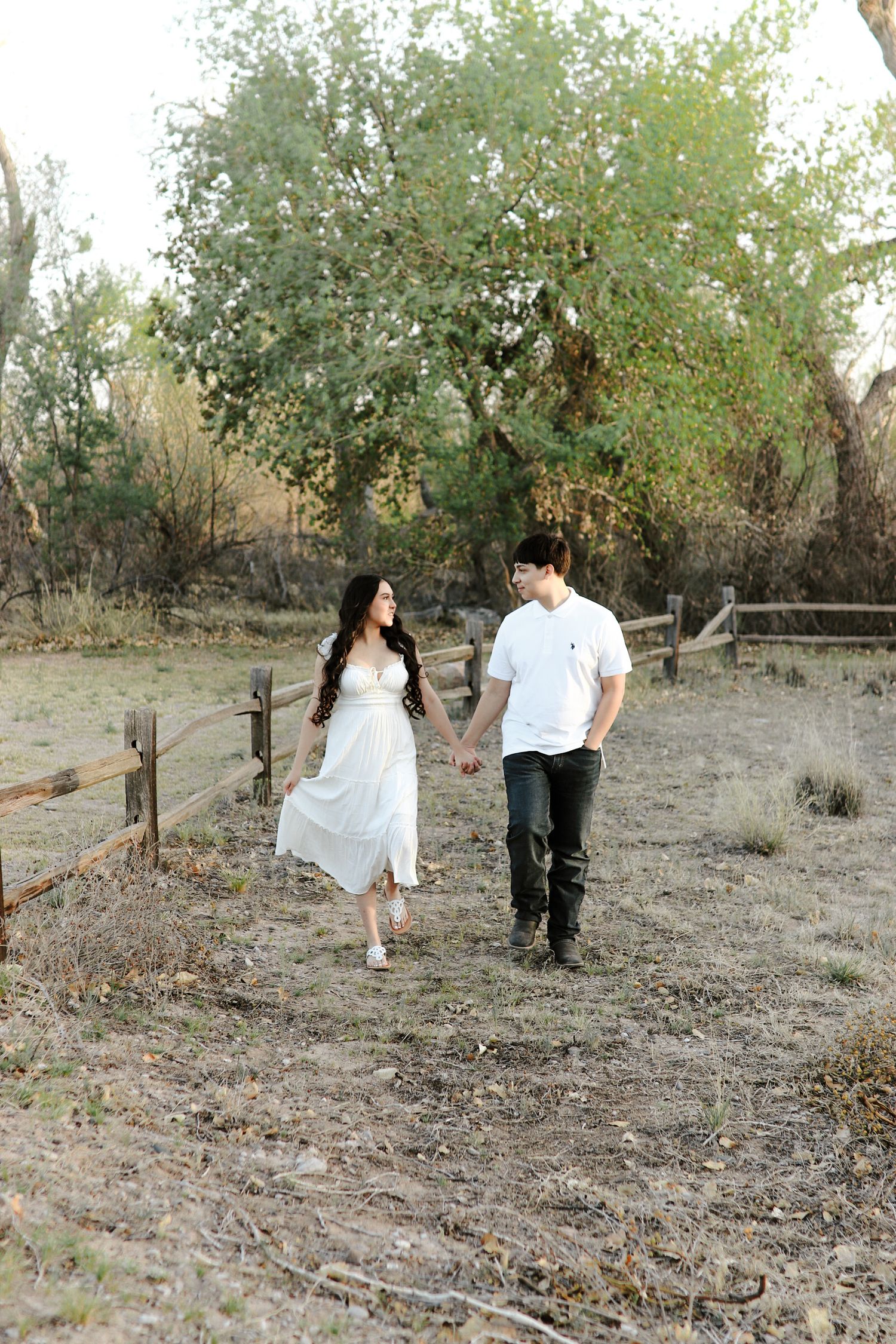 A couple holds hands and walks through a rustic outdoor setting with wooden fences and lush trees in the background.