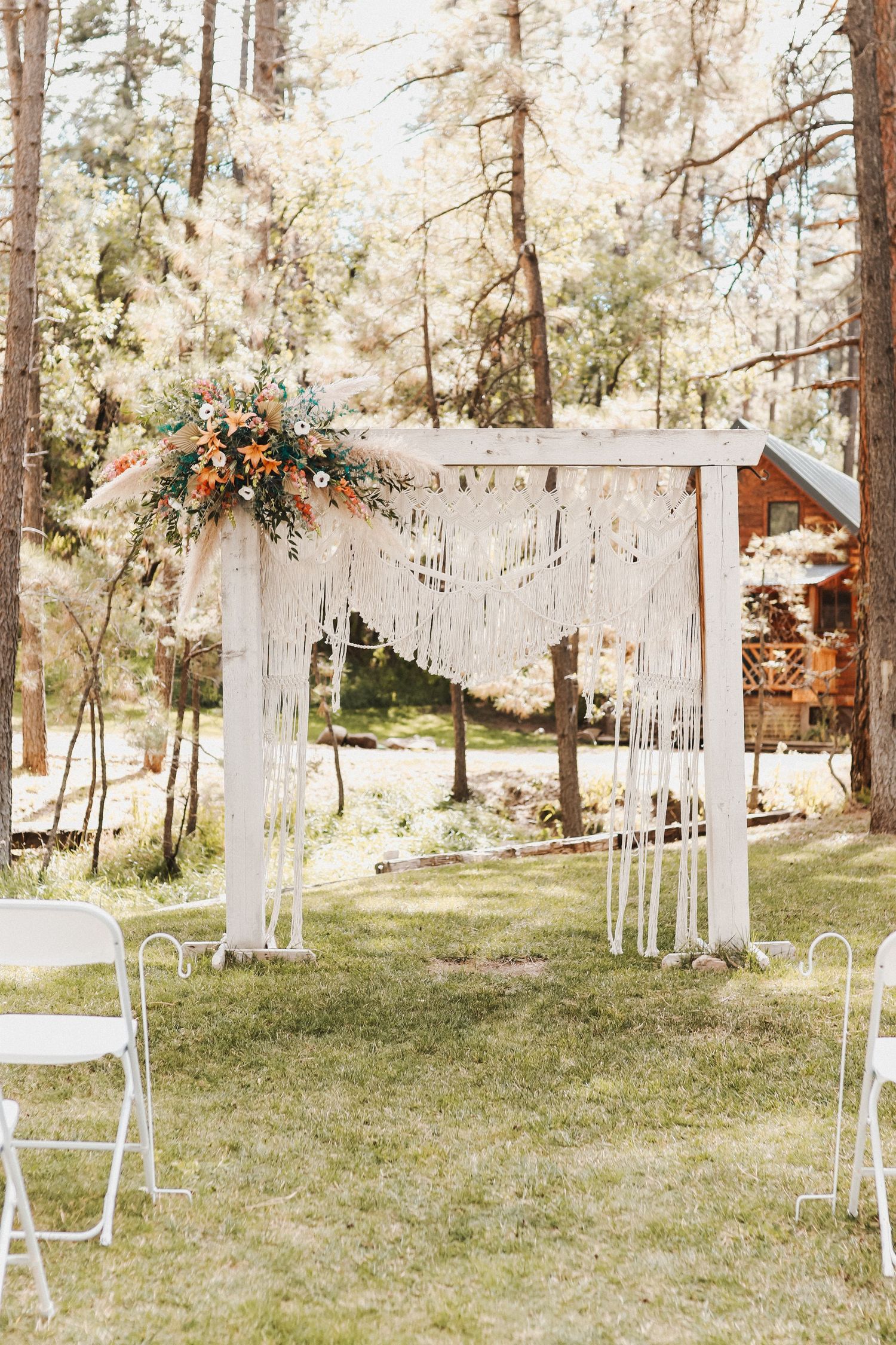 Outdoor wedding ceremony setup with white chairs and macrame arch in pine forest setting.
