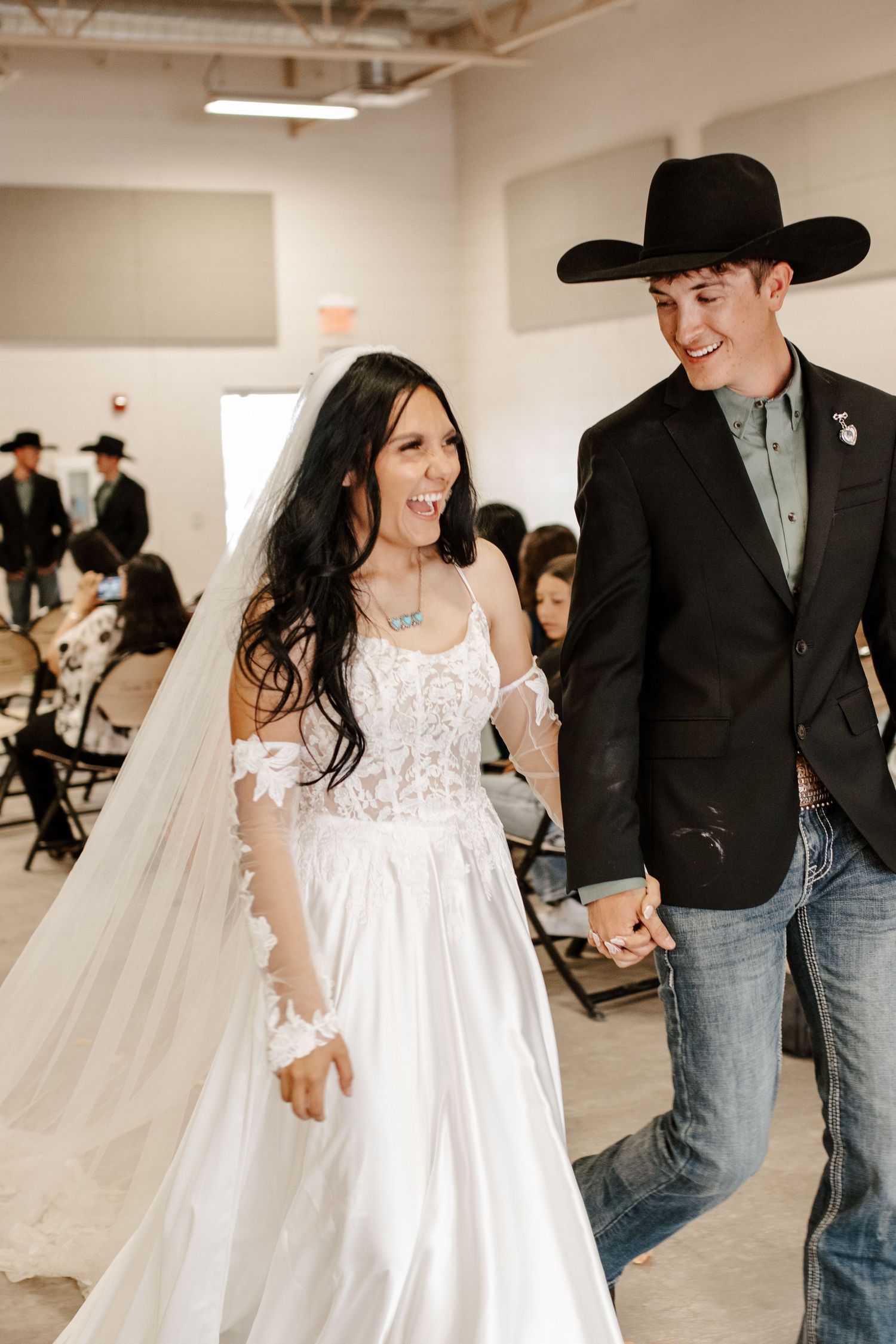 A bride in a white lace gown and veil walks hand-in-hand with a groom in a black cowboy hat at their wedding.