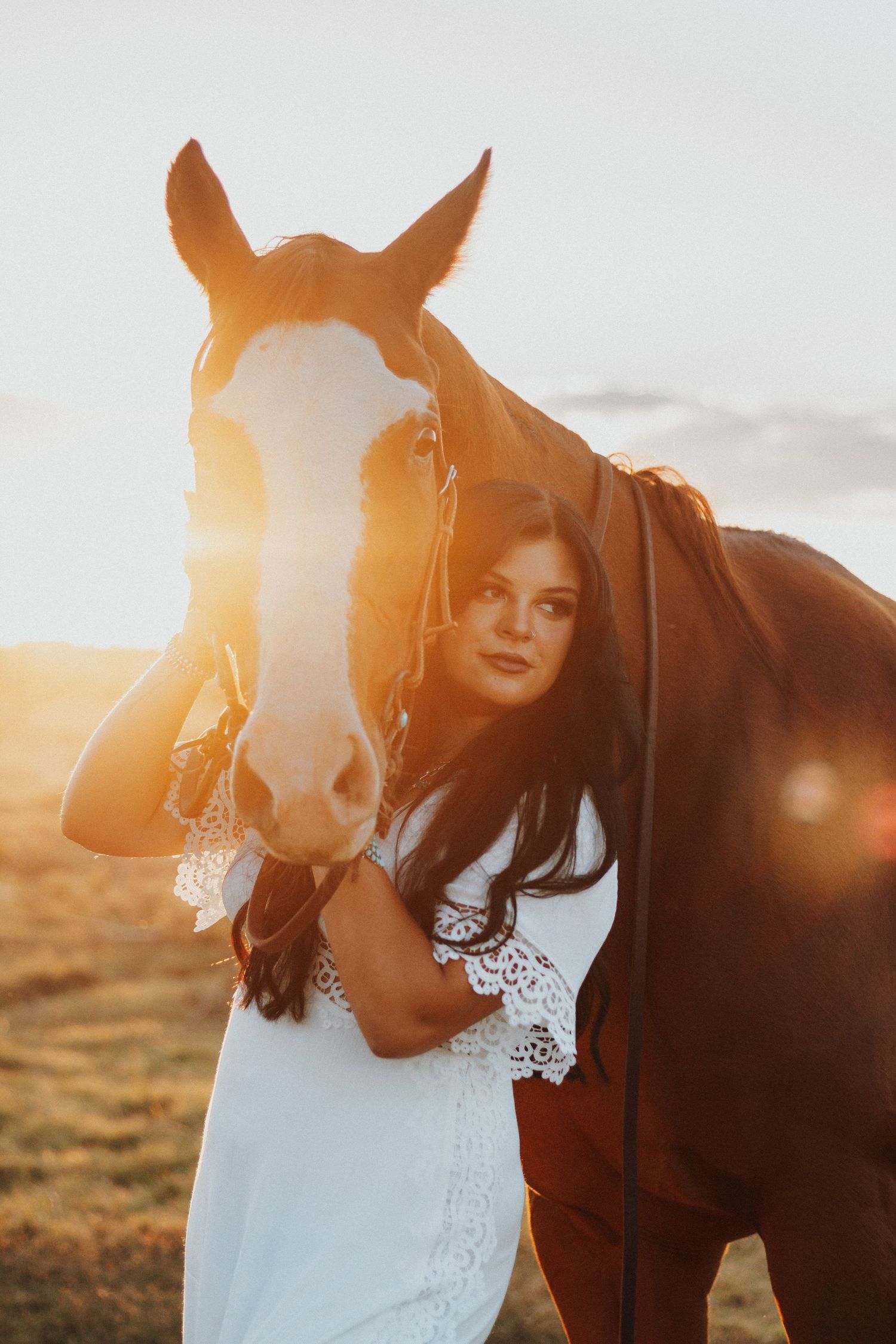 Golden sunset photo with brown horse and flowing white dress in scenic meadow setting.