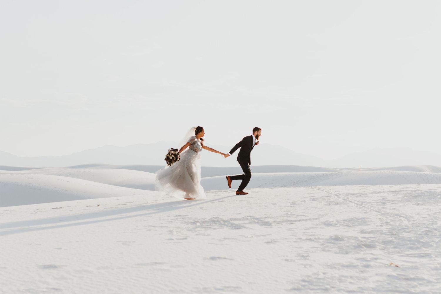 A couple runs playfully across pristine white sand dunes during a romantic photoshoot.