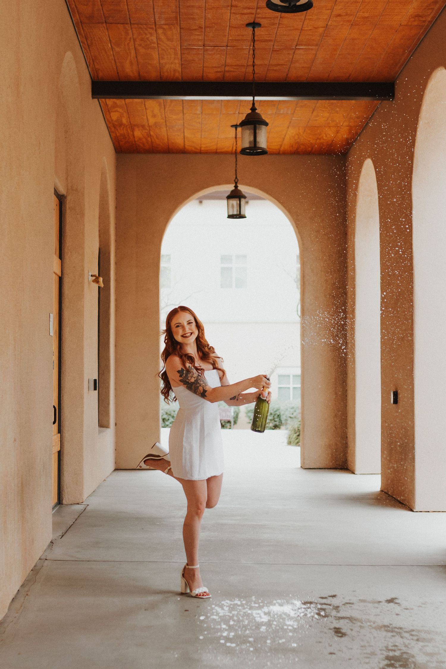 Graduate celebrating with champagne in white dress under Mediterranean-style archway.