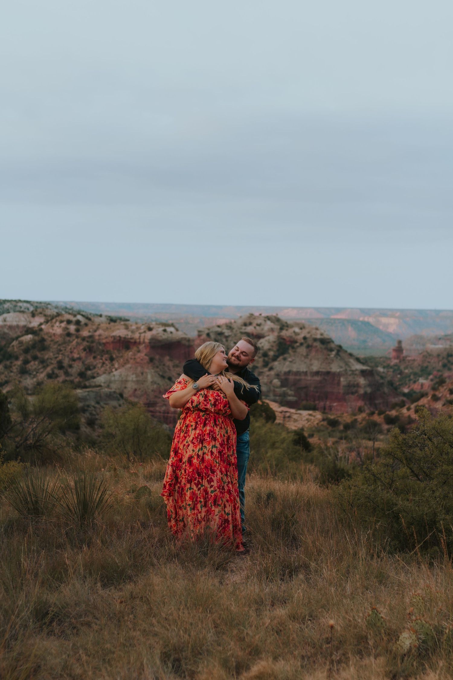 Couple embracing in a red floral dress at Palo Duro Canyon, with dramatic red rock formations in the background.