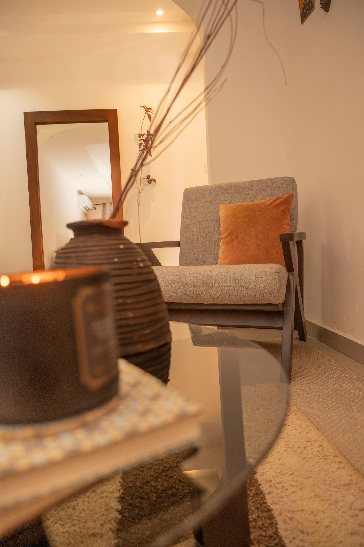 Close-up of grey armchair with orange pillow next to decorative vase and mirror in warm-lit room.