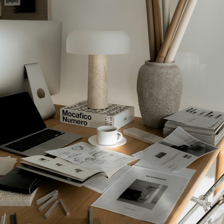 A minimalist wooden desk with a travertine lamp, open magazines, a coffee cup, and a laptop in a cozy home office.