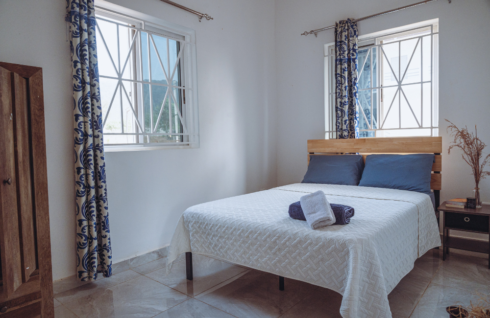 Cozy bedroom with white linens, navy pillows, and patterned curtains beside geometric windows.