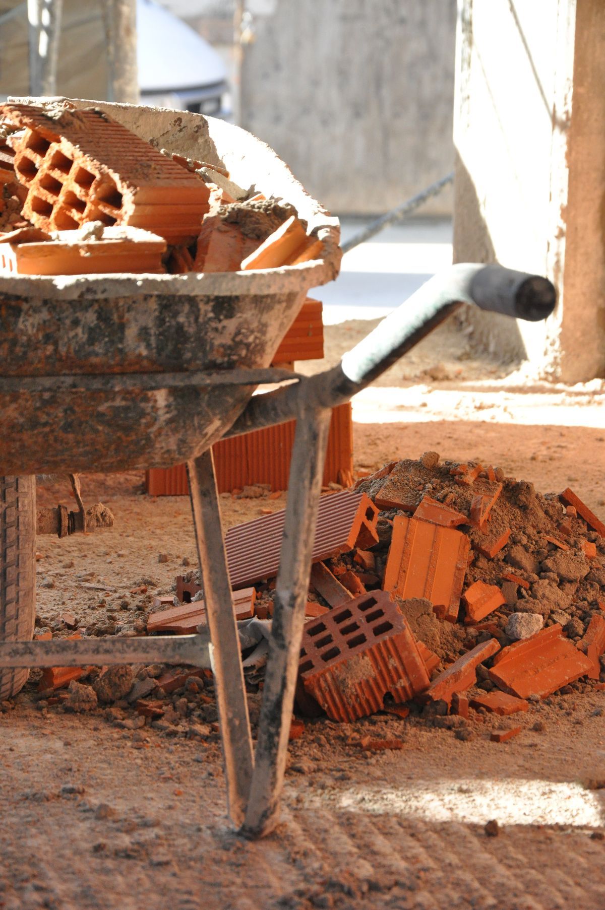 A wheelbarrow filled with red bricks and cement sits on a dusty construction site surrounded by broken bricks.