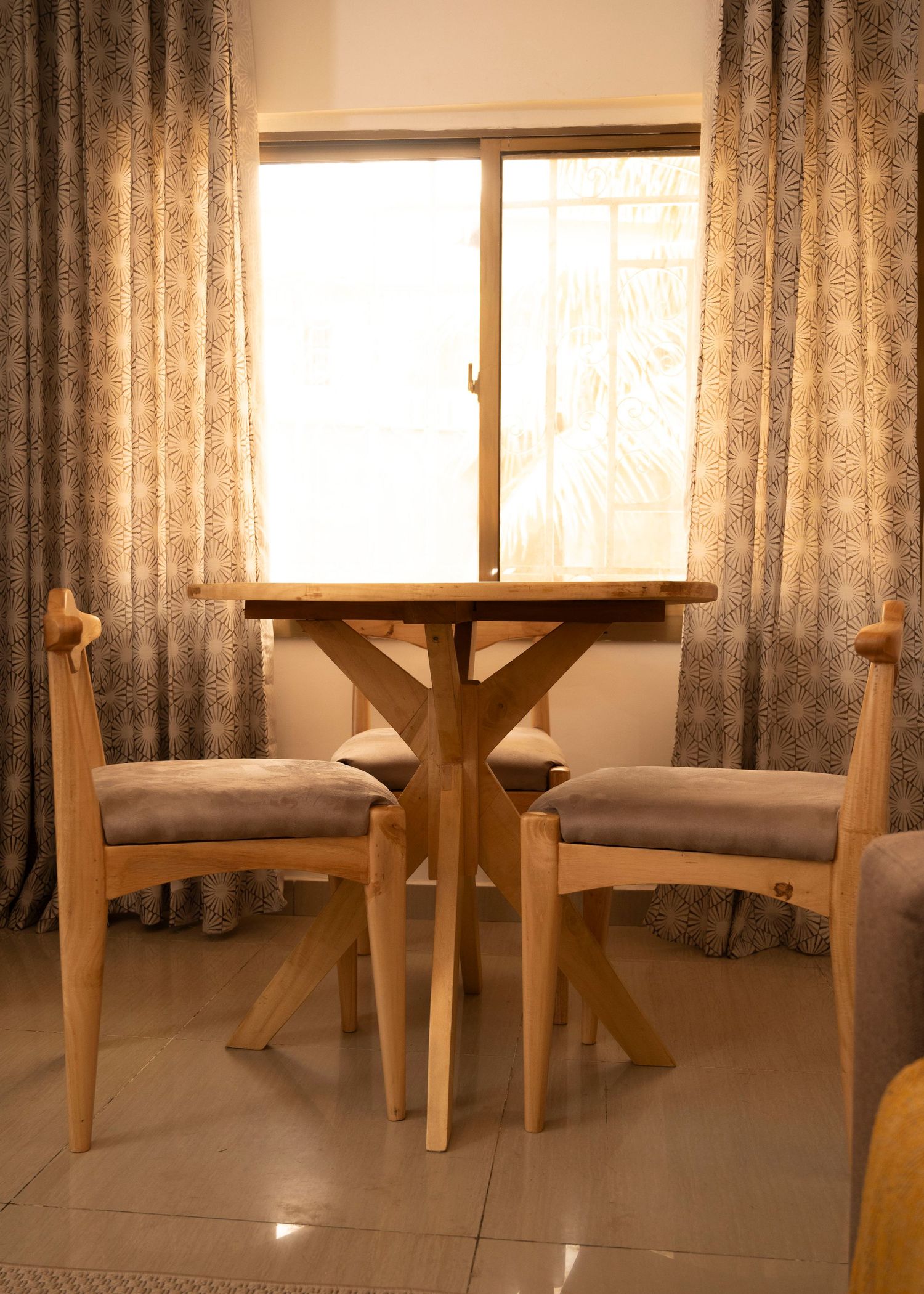 Dining nook with light wood chairs and X-base table framed by textured curtains and natural light.