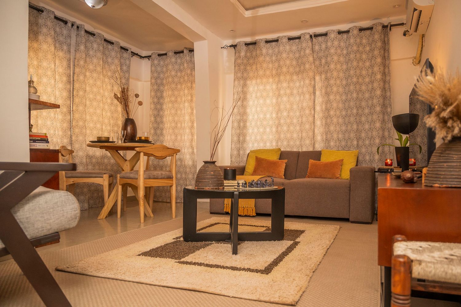 Cozy apartment interior featuring a grey couch, wooden furniture, and patterned curtains in warm lighting.