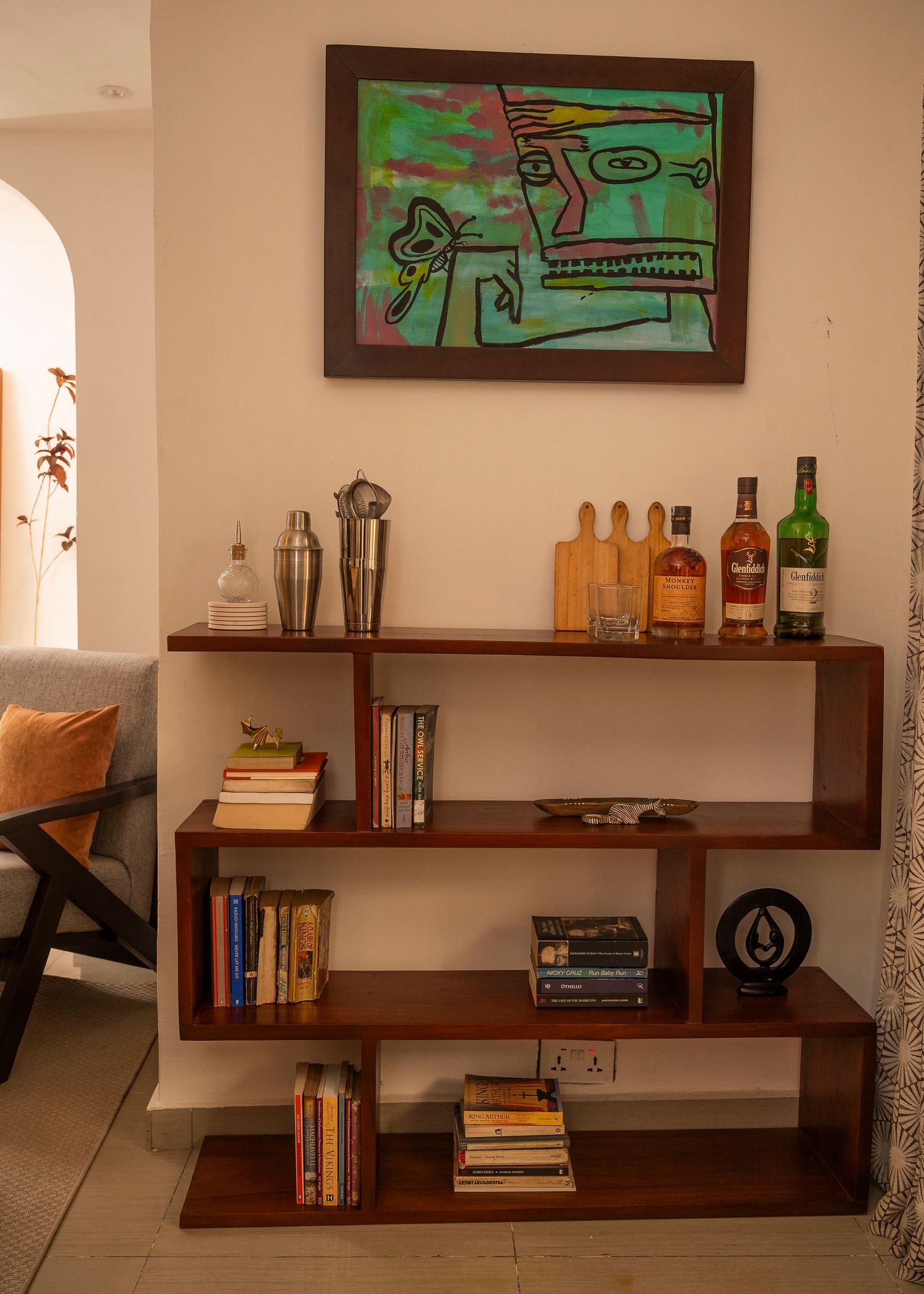 Modern bookshelf with geometric design displaying books, liquor bottles and vibrant green wall art.