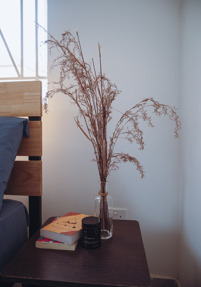 Dried floral arrangement in glass vase on dark wooden side table with books.