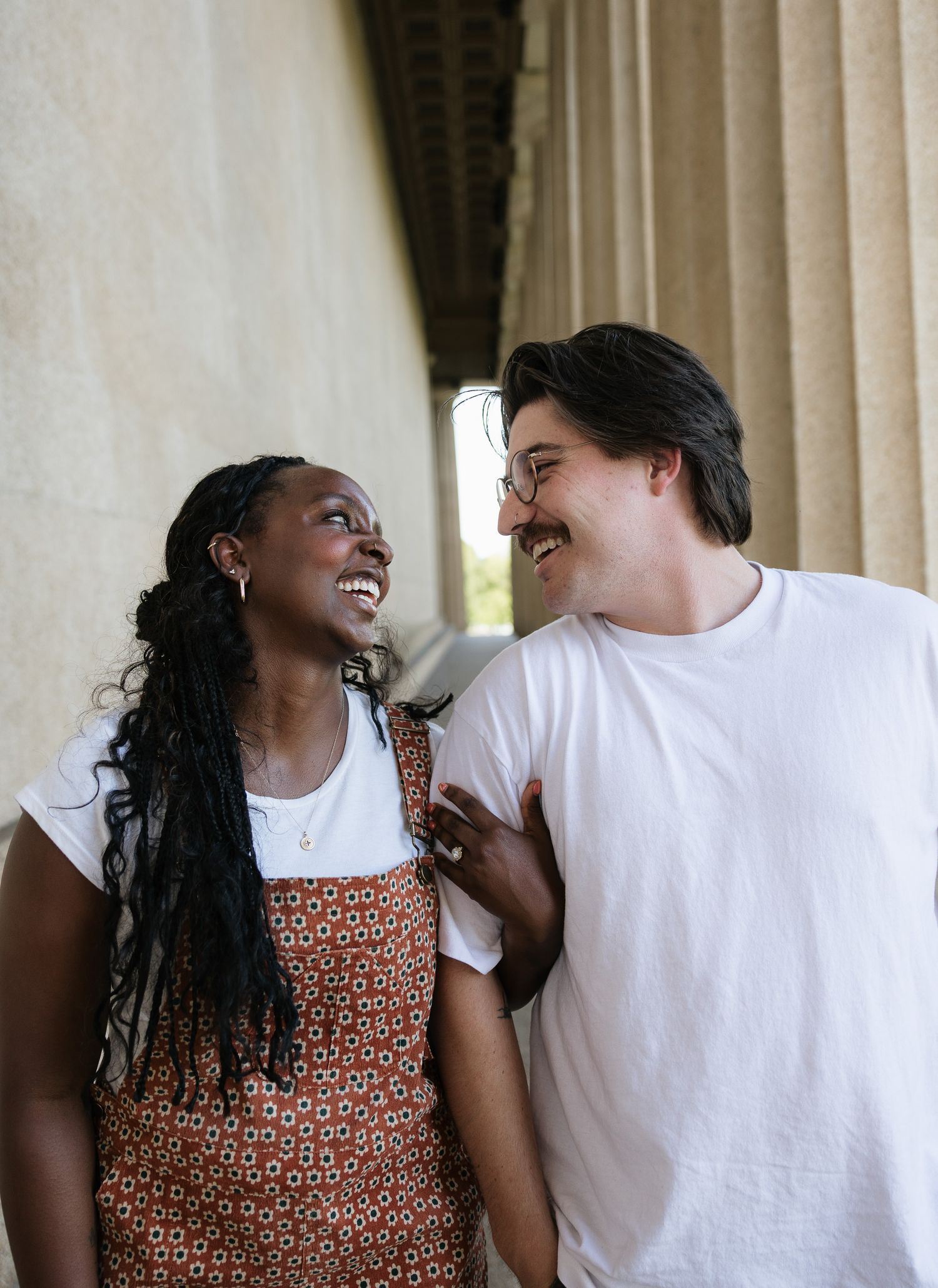 A joyful interracial couple smiling at each other beside tall stone columns outdoors.