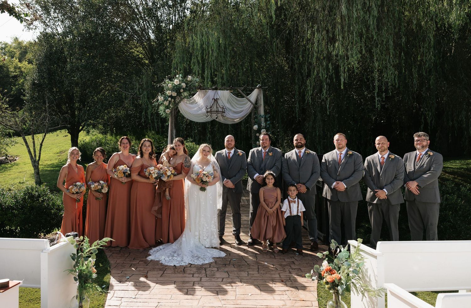 Wedding party poses outdoors under floral arch with bridesmaids in rust dresses and groomsmen in gray suits.