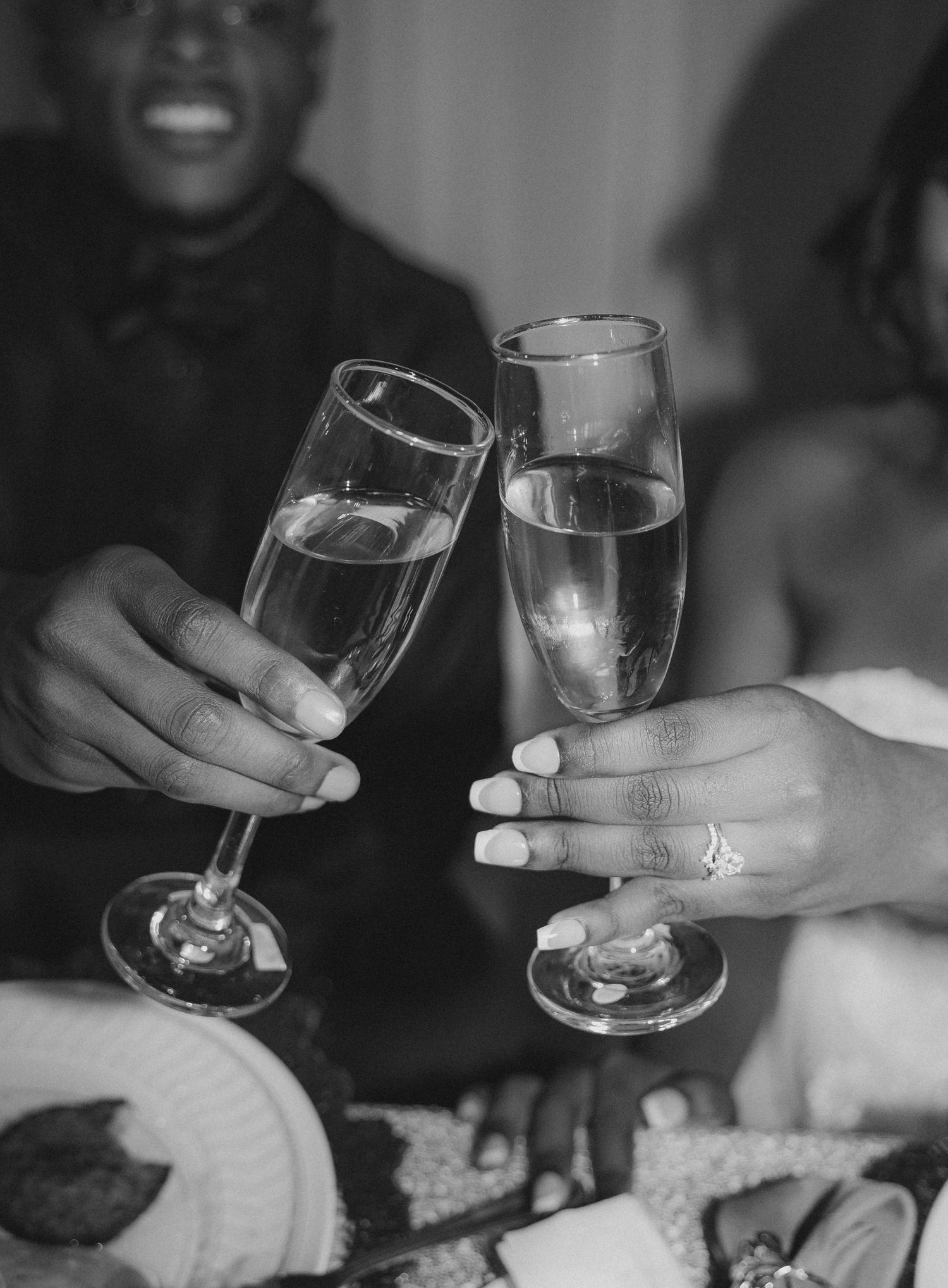 Black and white photo of two people clinking champagne flutes, with a diamond ring visible on one hand.