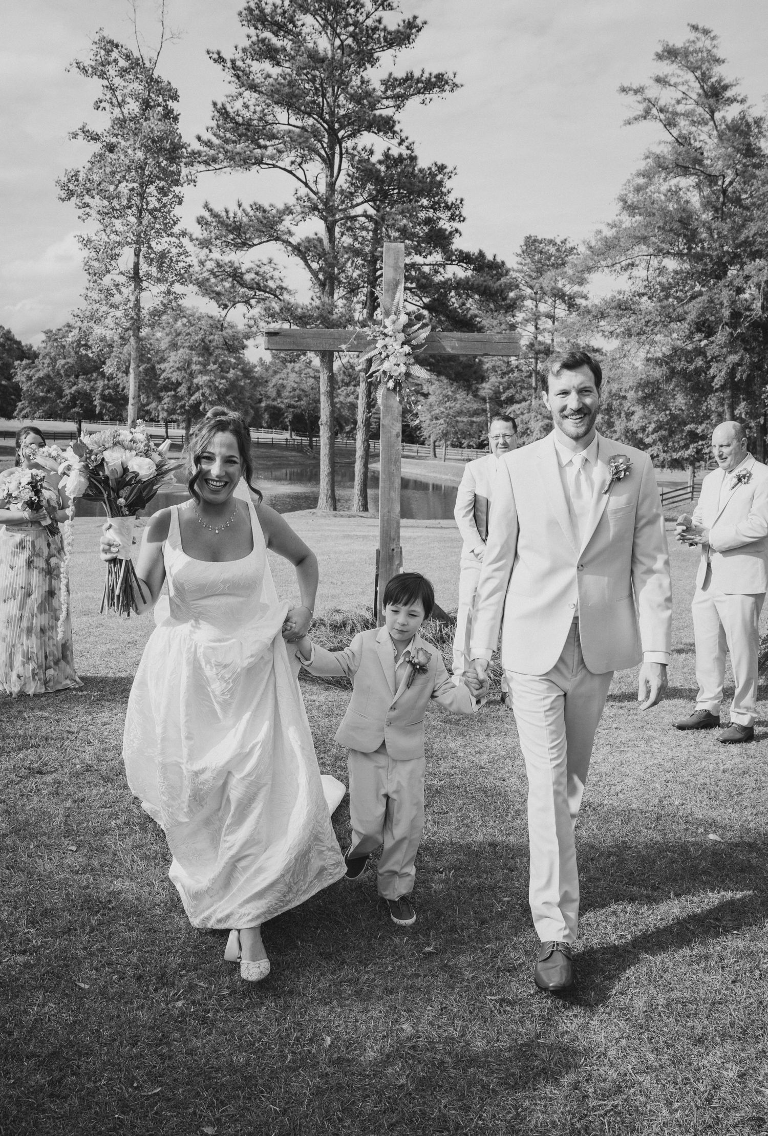 Bride and groom walk with young child after outdoor wedding ceremony in black and white photo.