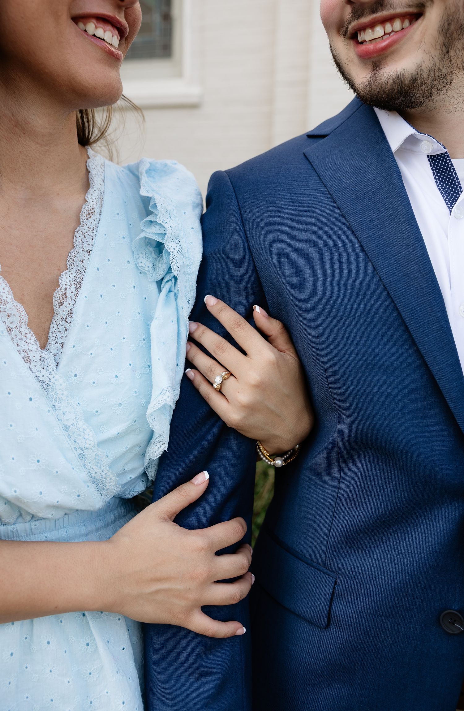 Smiling couple in formal wear, woman in light blue dress with gold ring holds mans arm in navy suit.