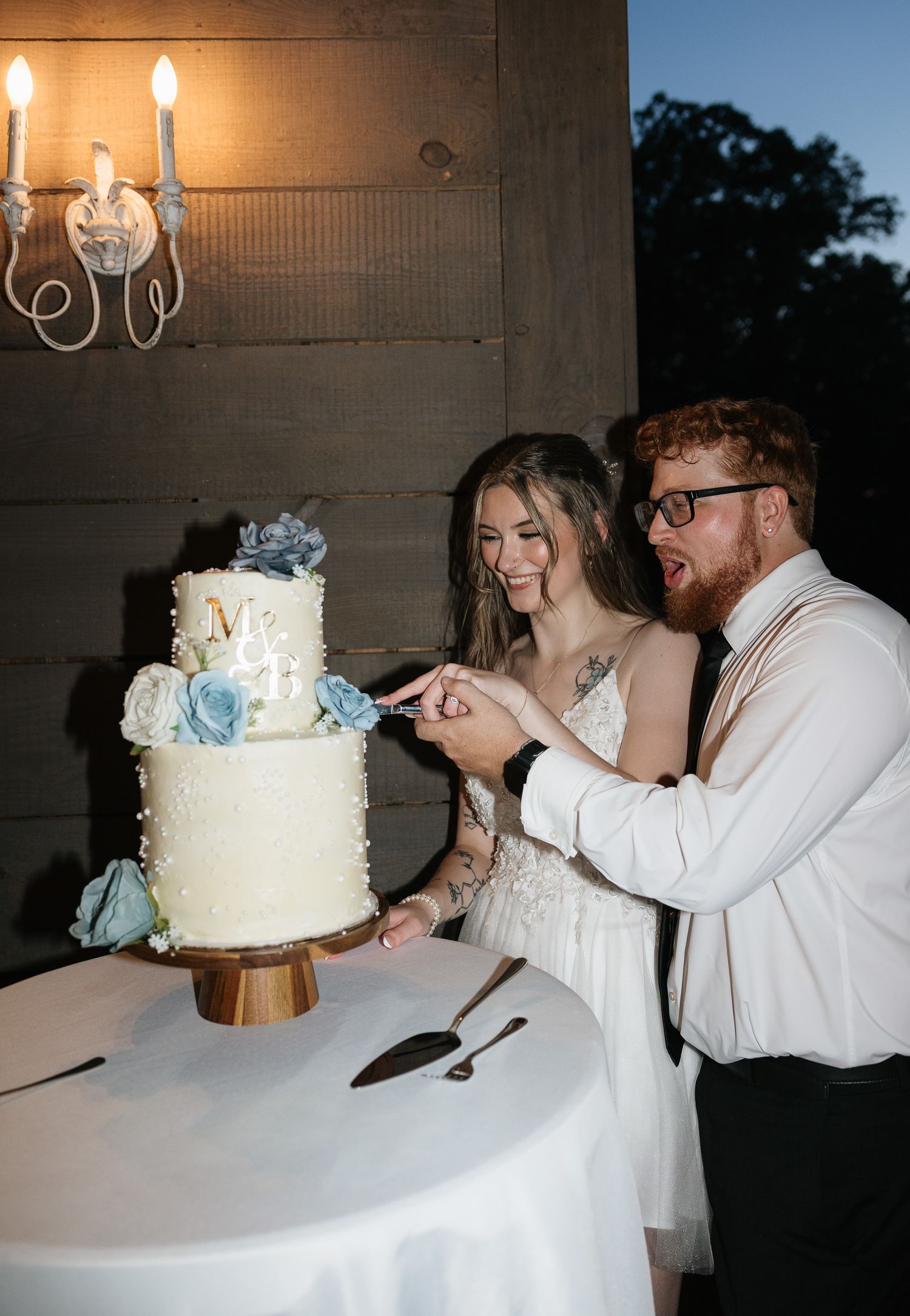 Smiling bride and groom cutting two-tier white wedding cake decorated with blue flowers at evening reception.