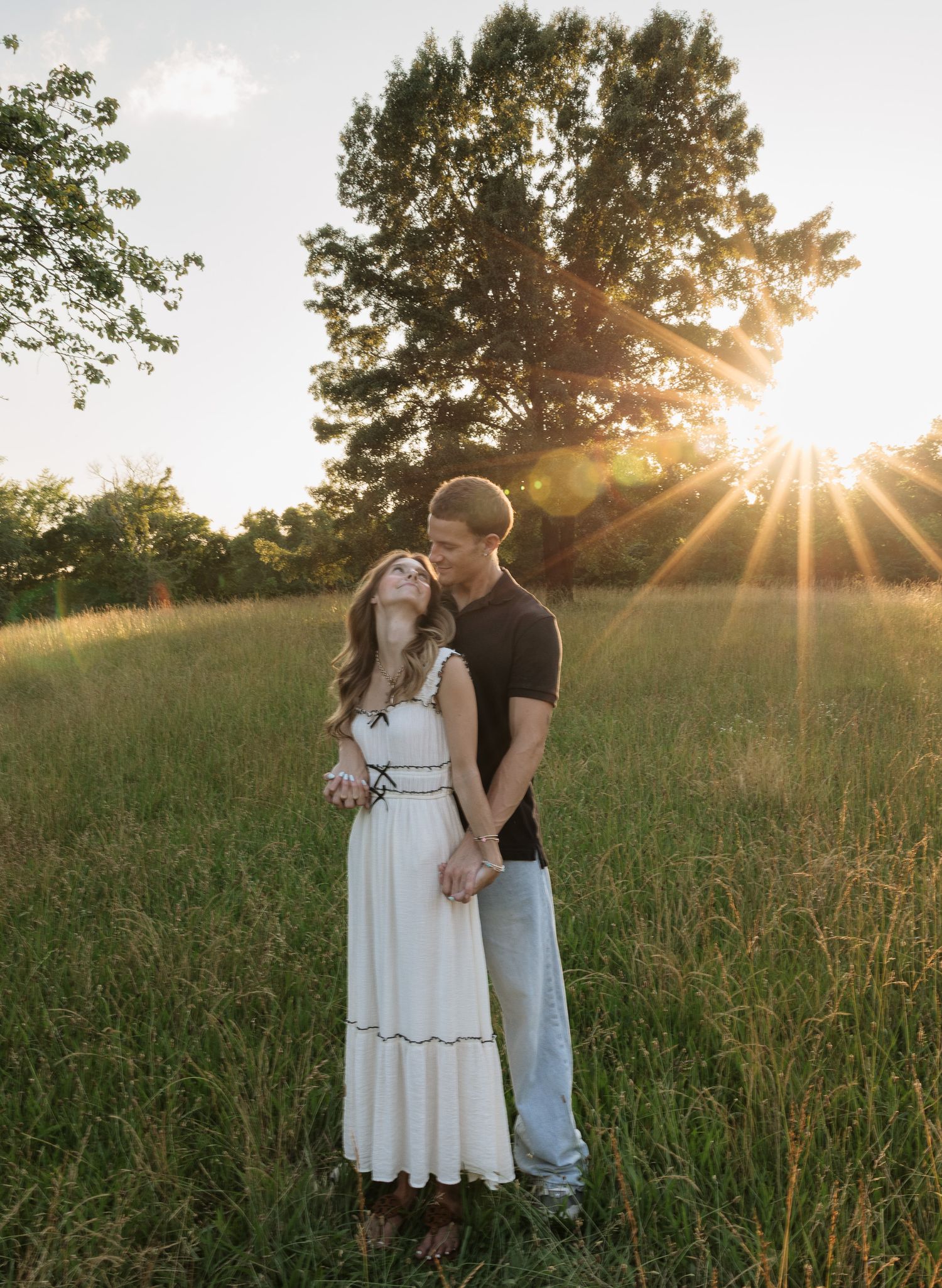 Couple embracing in golden sunlit meadow at sunset, woman in white dress, man in black shirt, large tree background.