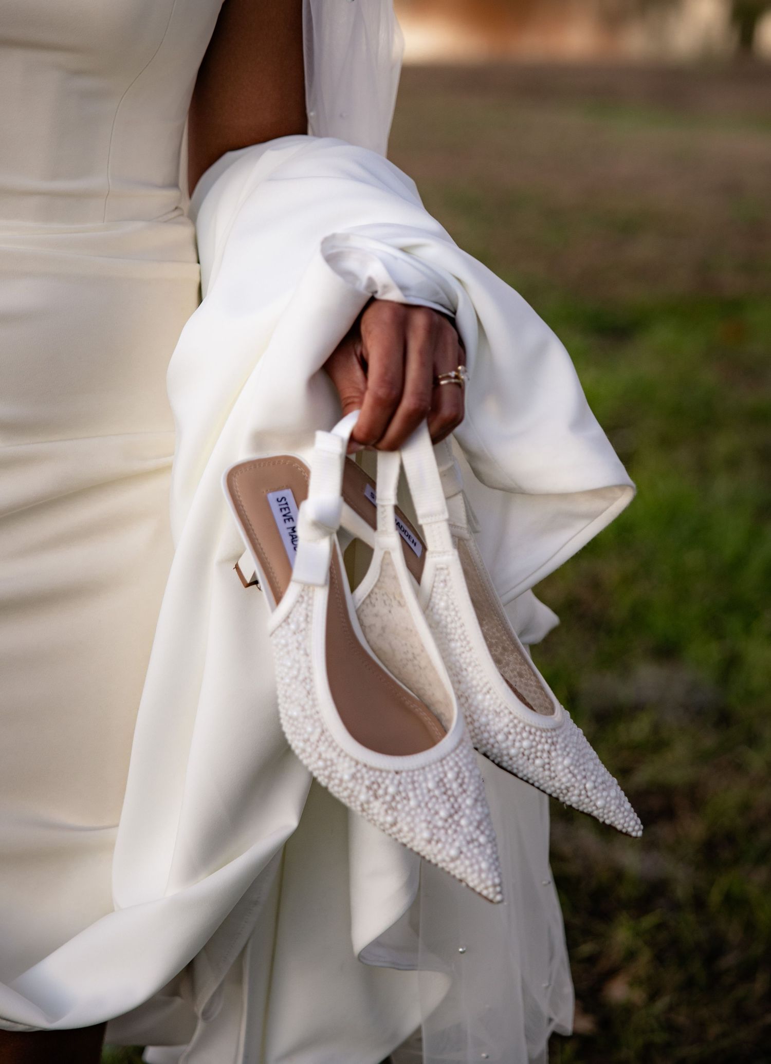 Elegant white bridal shoes with sparkly crystal embellishments held against flowing white dress fabric in outdoor setting.