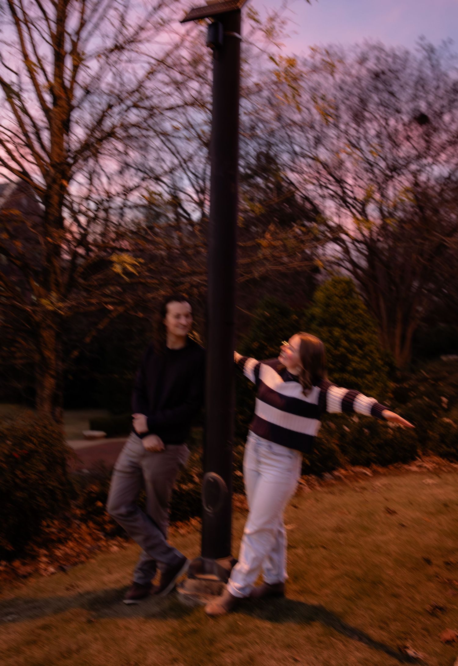 Two young people posing playfully around a pole in an autumn park at dusk.
