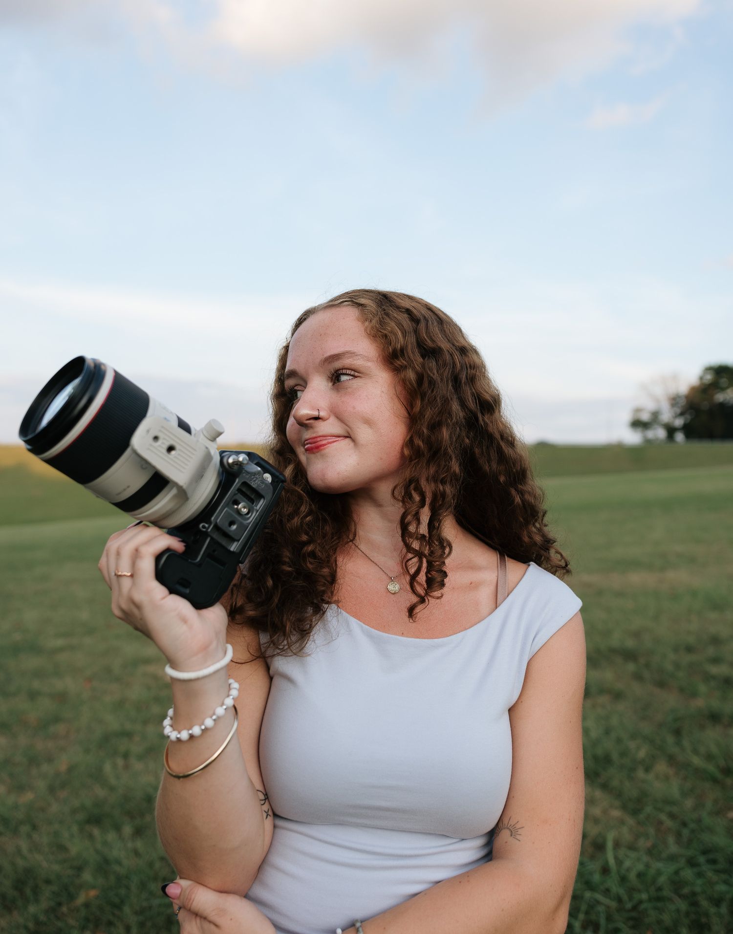 Young woman with curly hair holding a Canon camera with telephoto lens outdoors on a green field at dusk.