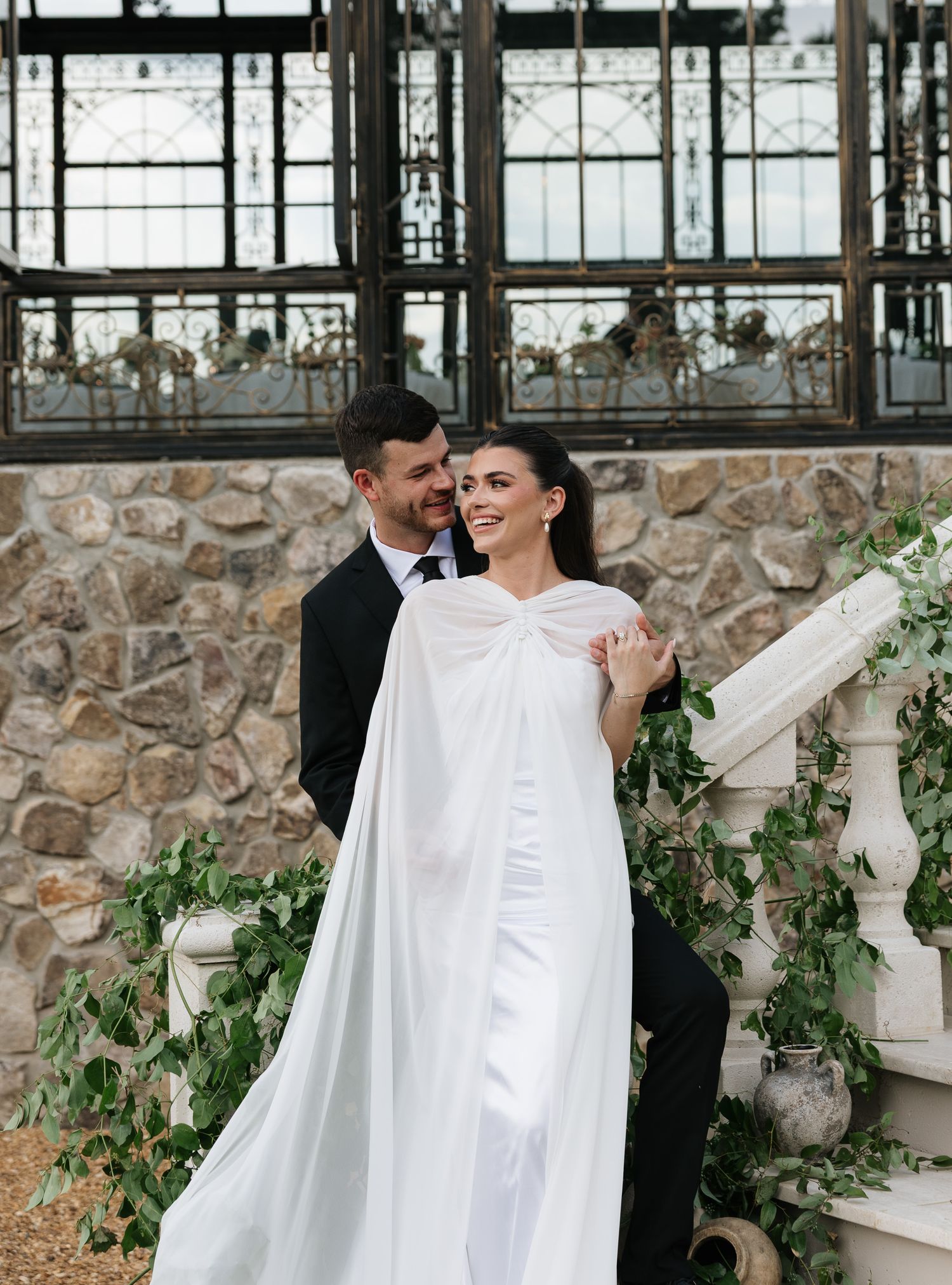 Elegant bride in white cape gown and groom in black tuxedo pose on stone staircase adorned with greenery.