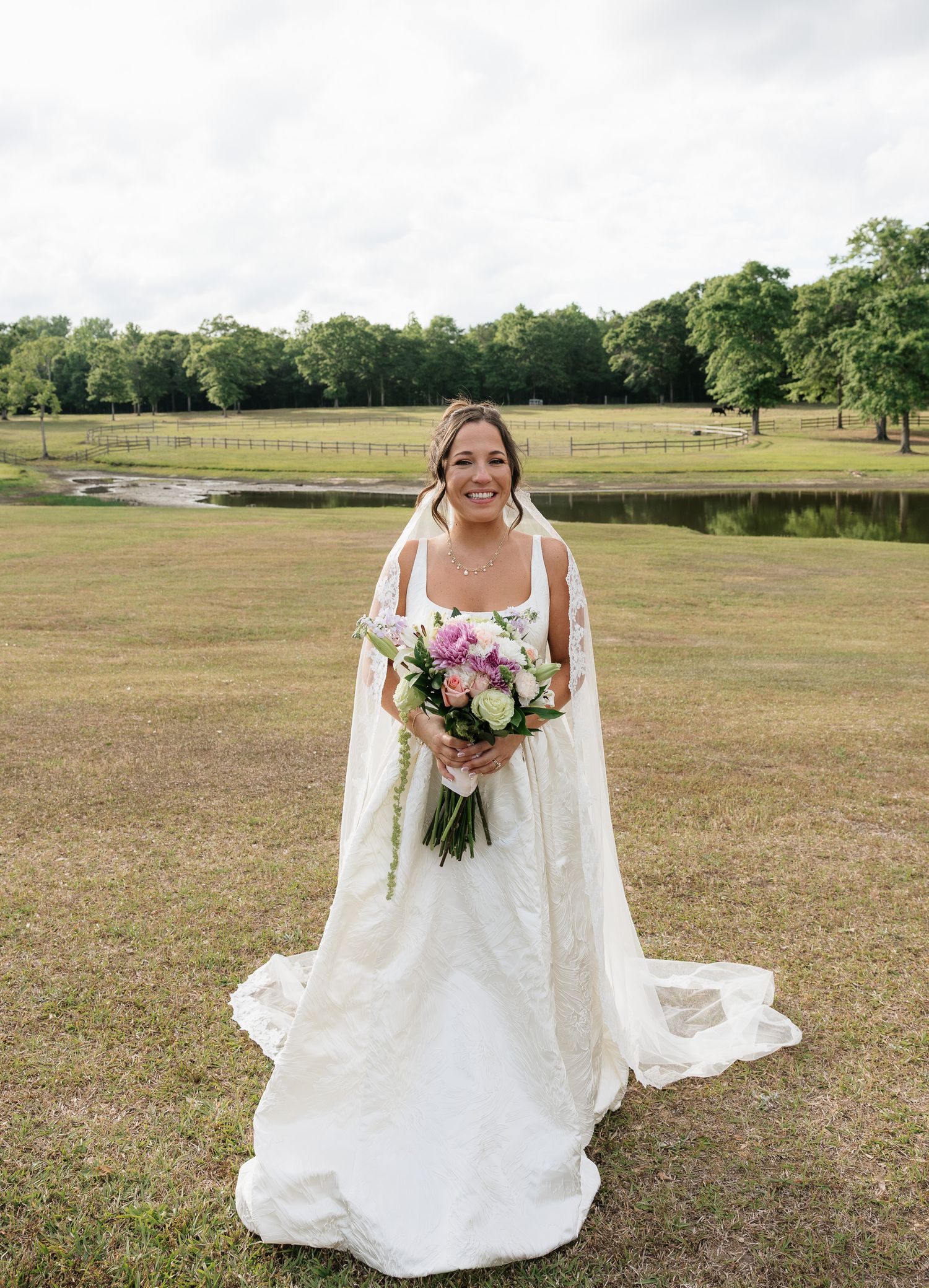 Bride in white gown with cape holds pink bouquet on green lawn with pond and trees backdrop.