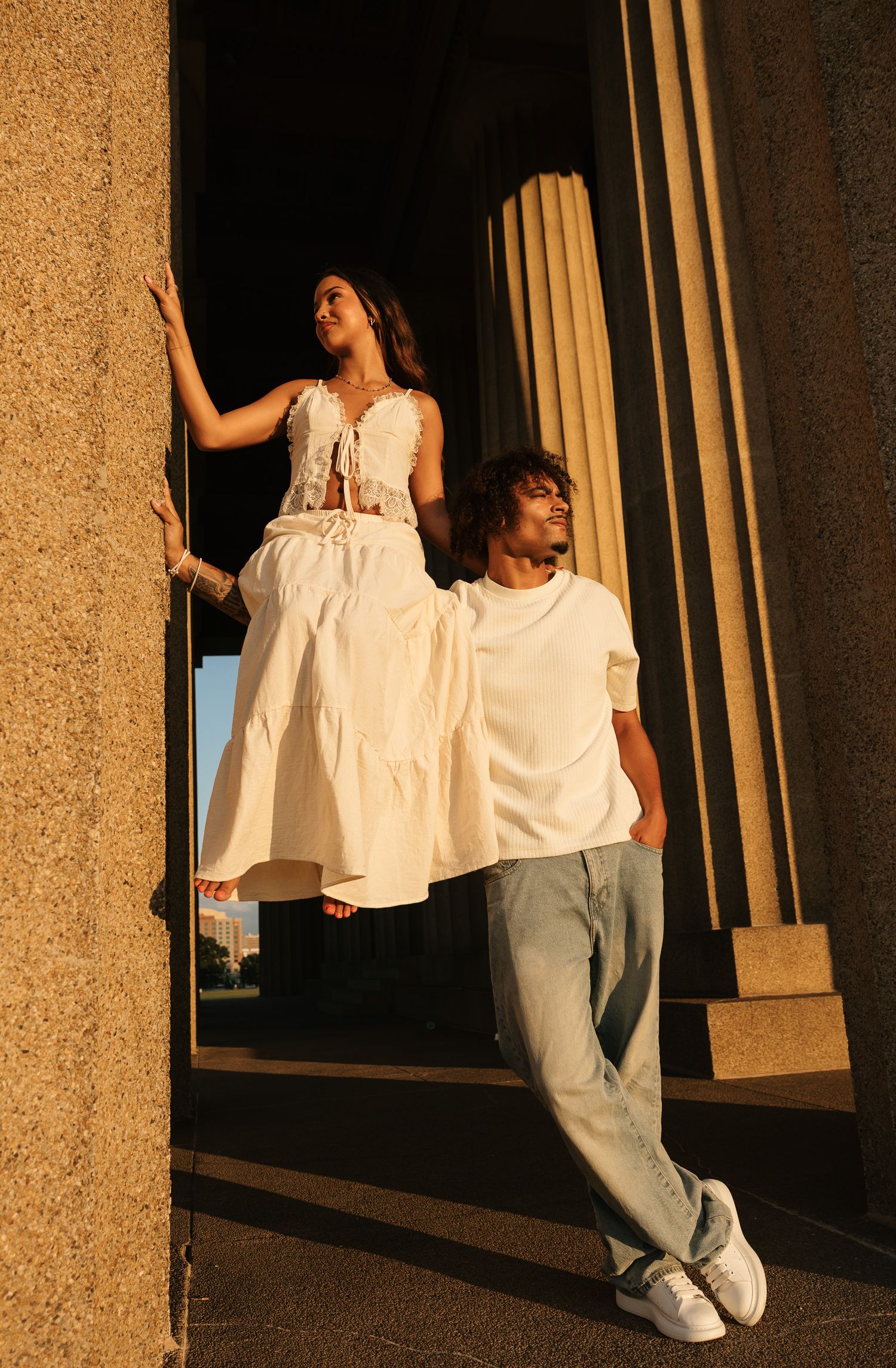 A woman in white dress and man in white tee and jeans pose between large stone columns in golden hour light.